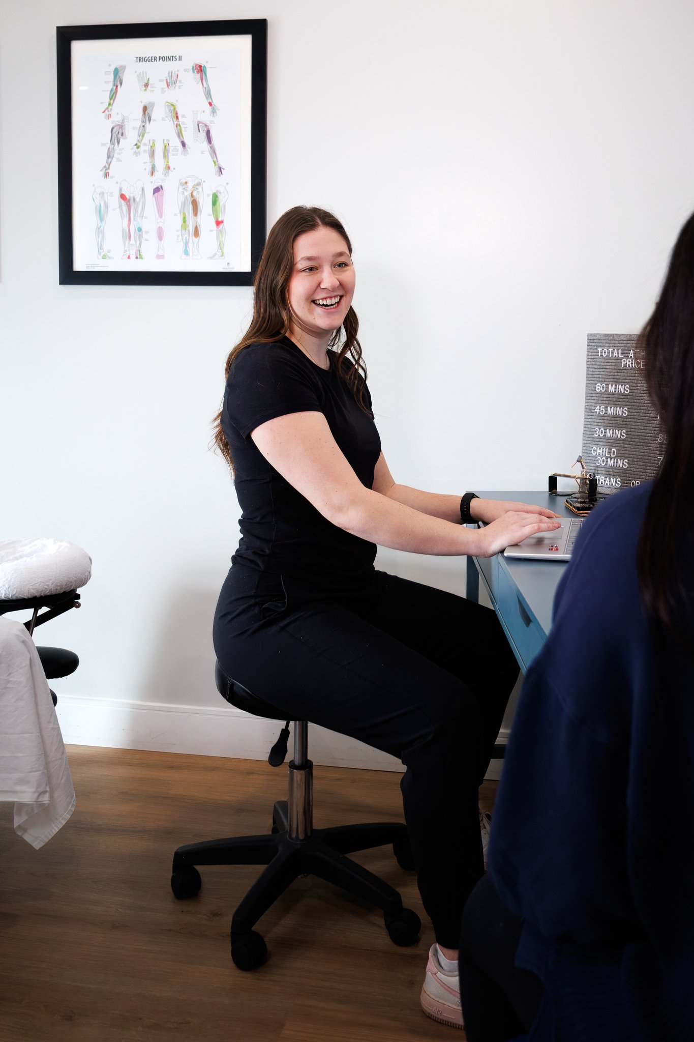 A woman with long brown hair sitting at a desk, laughing, with a clock and a poster of trigger points on the wall behind her.