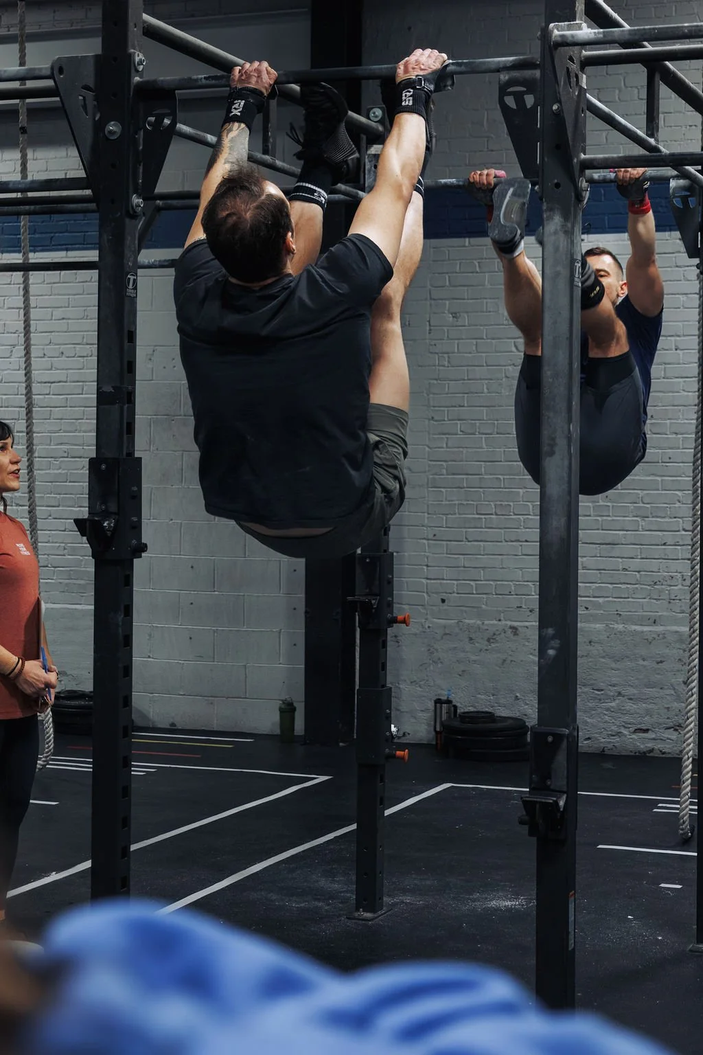 Two men are doing pull-ups on a pull-up bar in a gym, with a woman observing nearby.