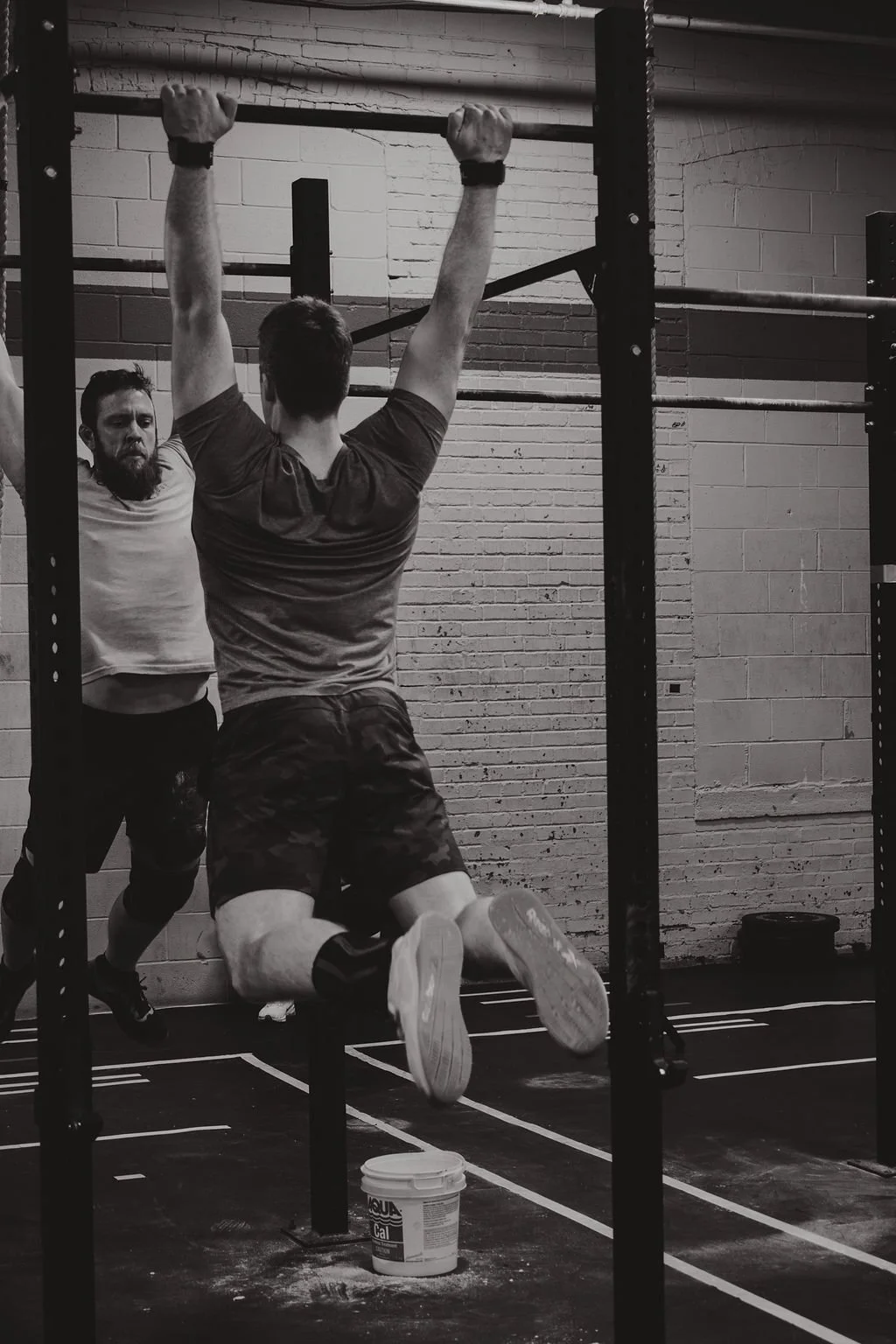 A man is doing pull-ups on a pull-up bar in a gym while another man is standing nearby observing.