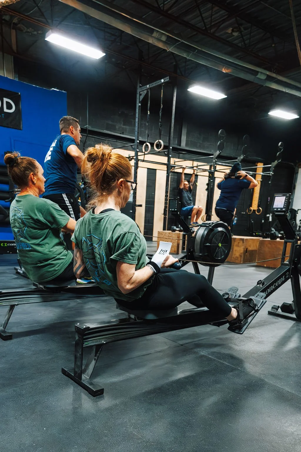People working out in a crossfit gym, with women using rowing machines and others doing pull-ups and hang exercises on pull-up bars.