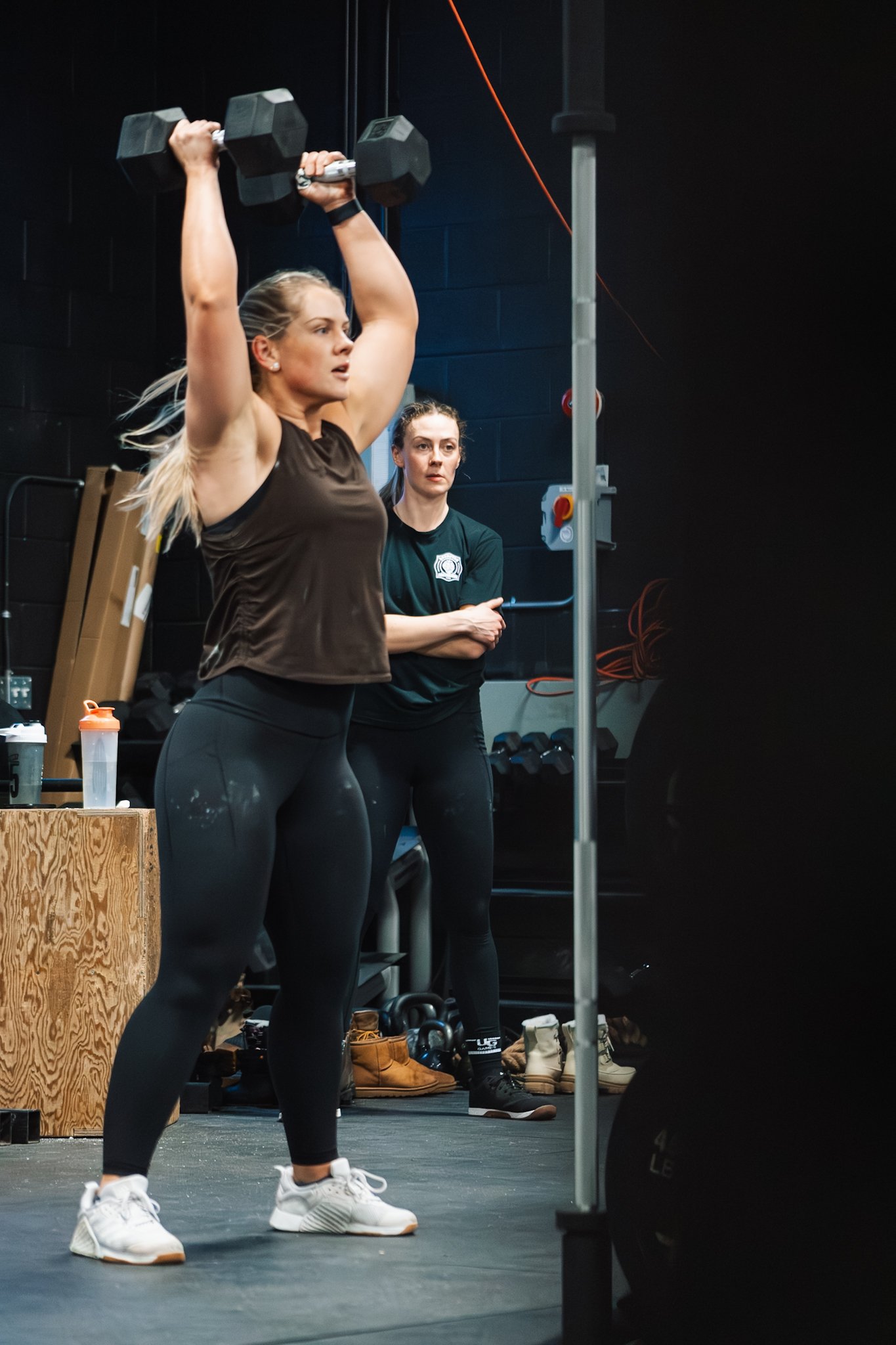 A woman lifting a dumbbell overhead in a gym with another woman watching.