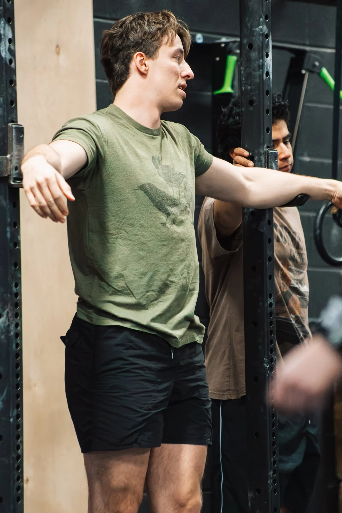 A man with light skin and brown hair stretching at a gym, wearing a green t-shirt with a bird graphic and black shorts, standing next to a black metal squat rack.