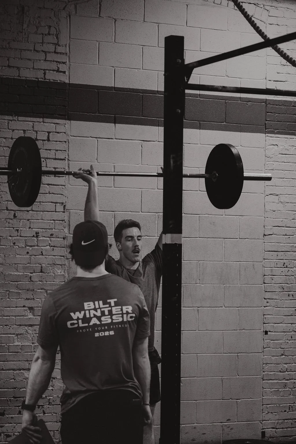 Two men in a gym lifting a barbell overhead, one facing away in a Nike cap and t-shirt, and the other facing forward with a focused expression, beside a brick wall.