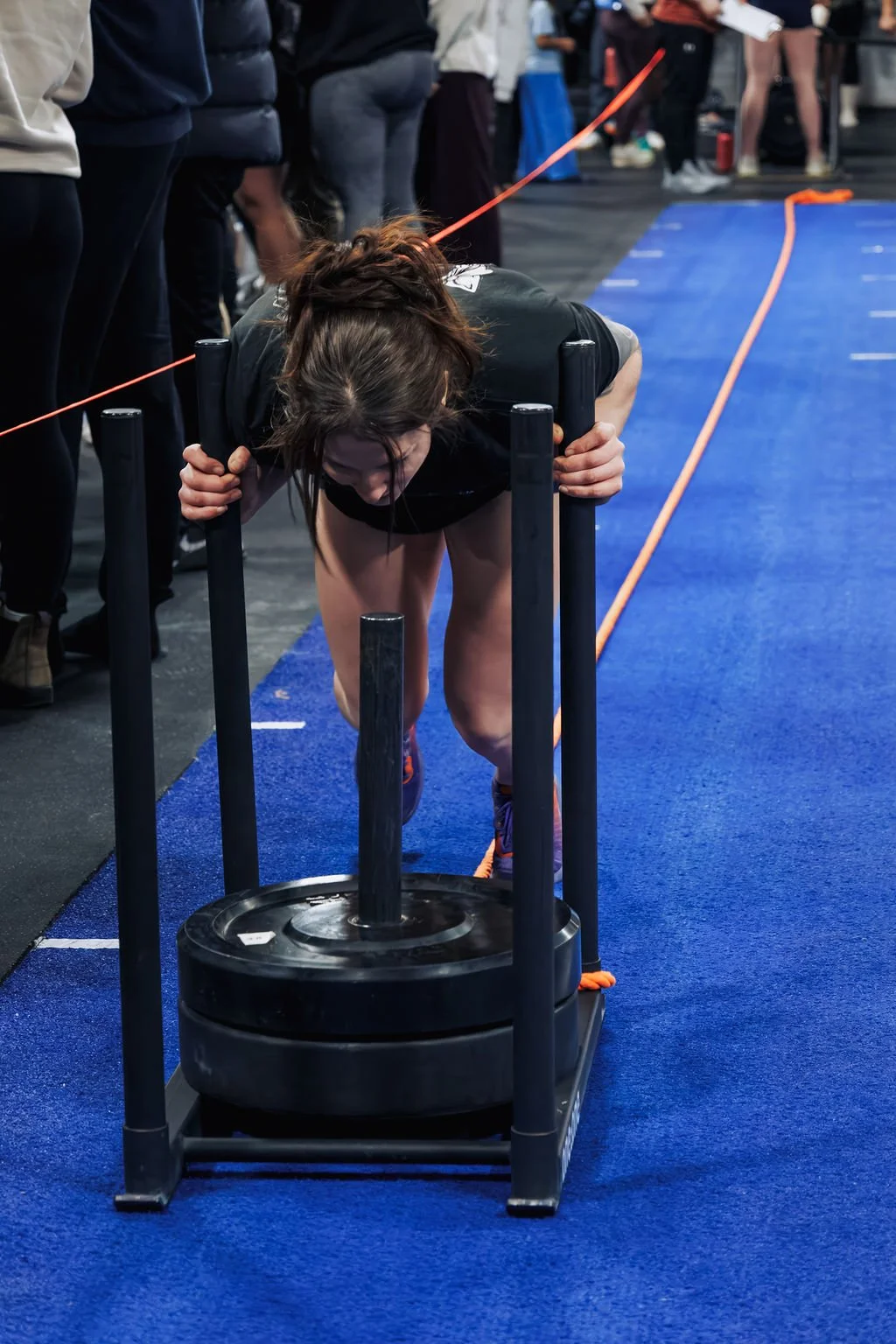 A woman pushing a sled with weights during her workout at a gym, with a crowd of people in the background.