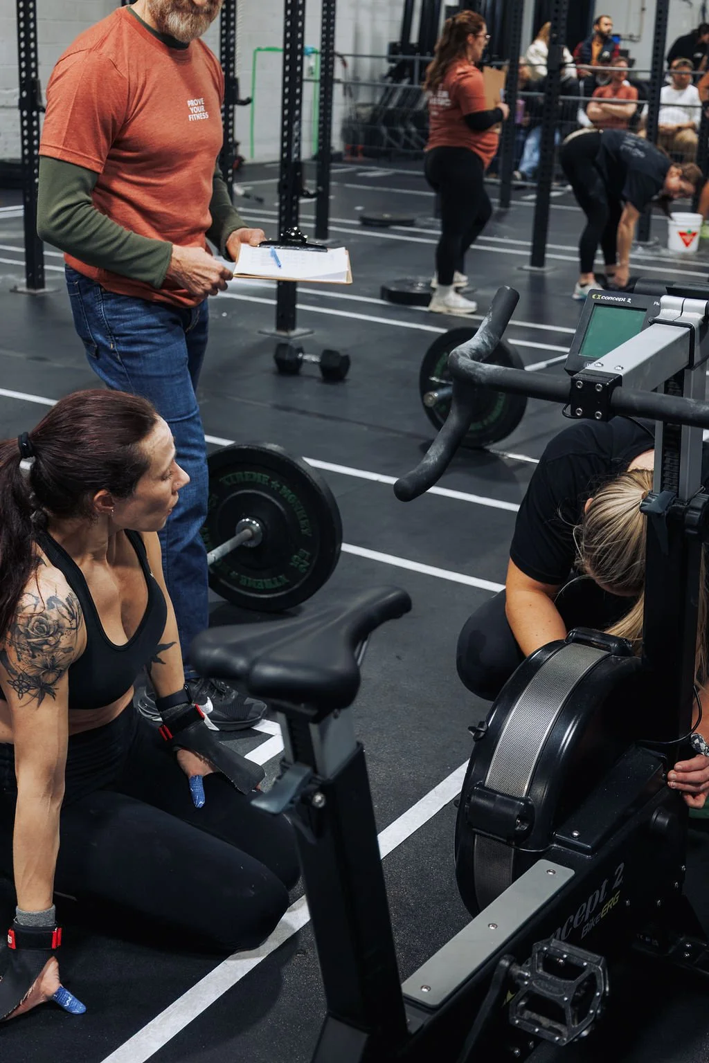 People preparing for a workout at a gym with a coach holding a clipboard, surrounded by fitness equipment, and other individuals exercising or waiting in the background.