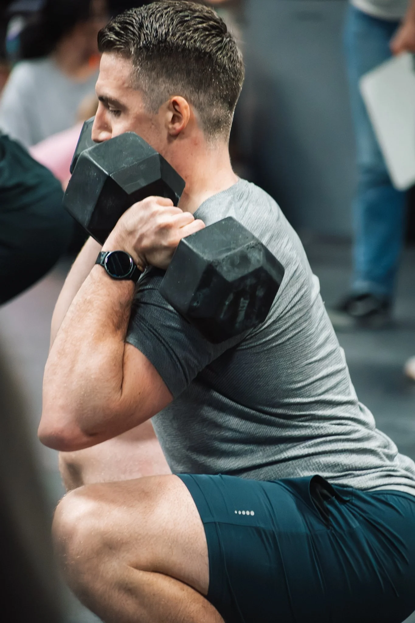A man wearing a gray shirt and black shorts performing a squat exercise while holding a dumbbell in each hand at a gym.