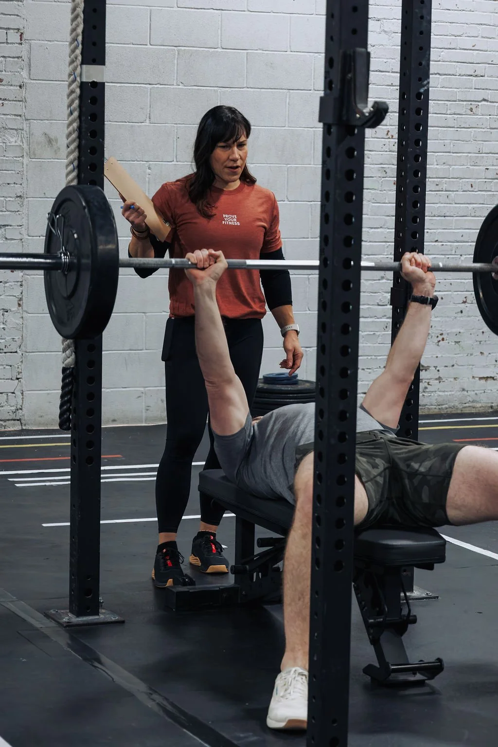 A woman spotter instructs a man performing a bench press at a gym, with weights on the barbell and gym equipment in the background.