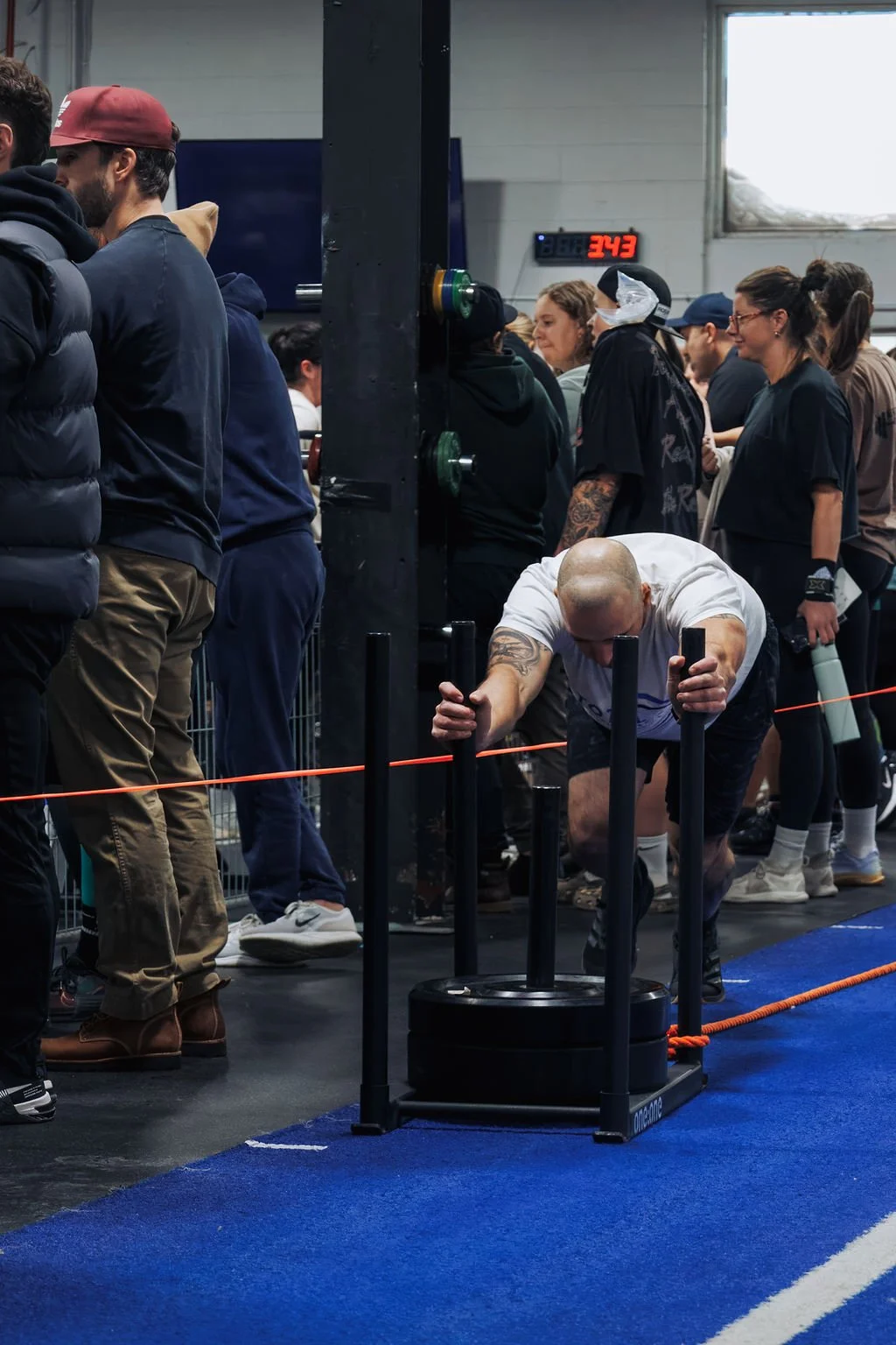 A man with a shaved head and tattoos on his arms is participating in a strongman competition, pushing a large weight sled on a blue carpeted surface, while spectators and other competitors stand nearby in a gym or indoor event space.