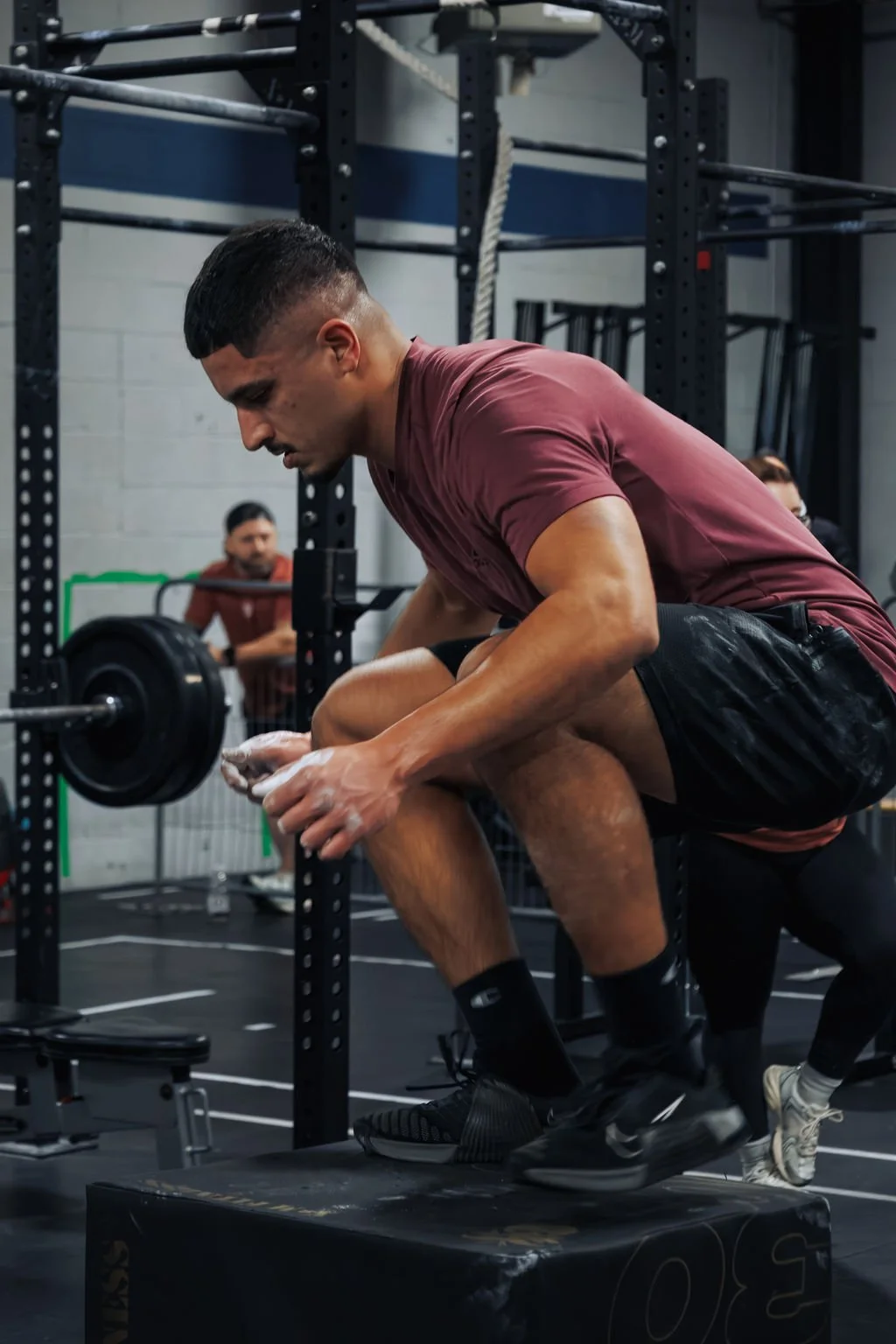 Man in maroon shirt performing a squat with a barbell in a gym, with an observer in the background.