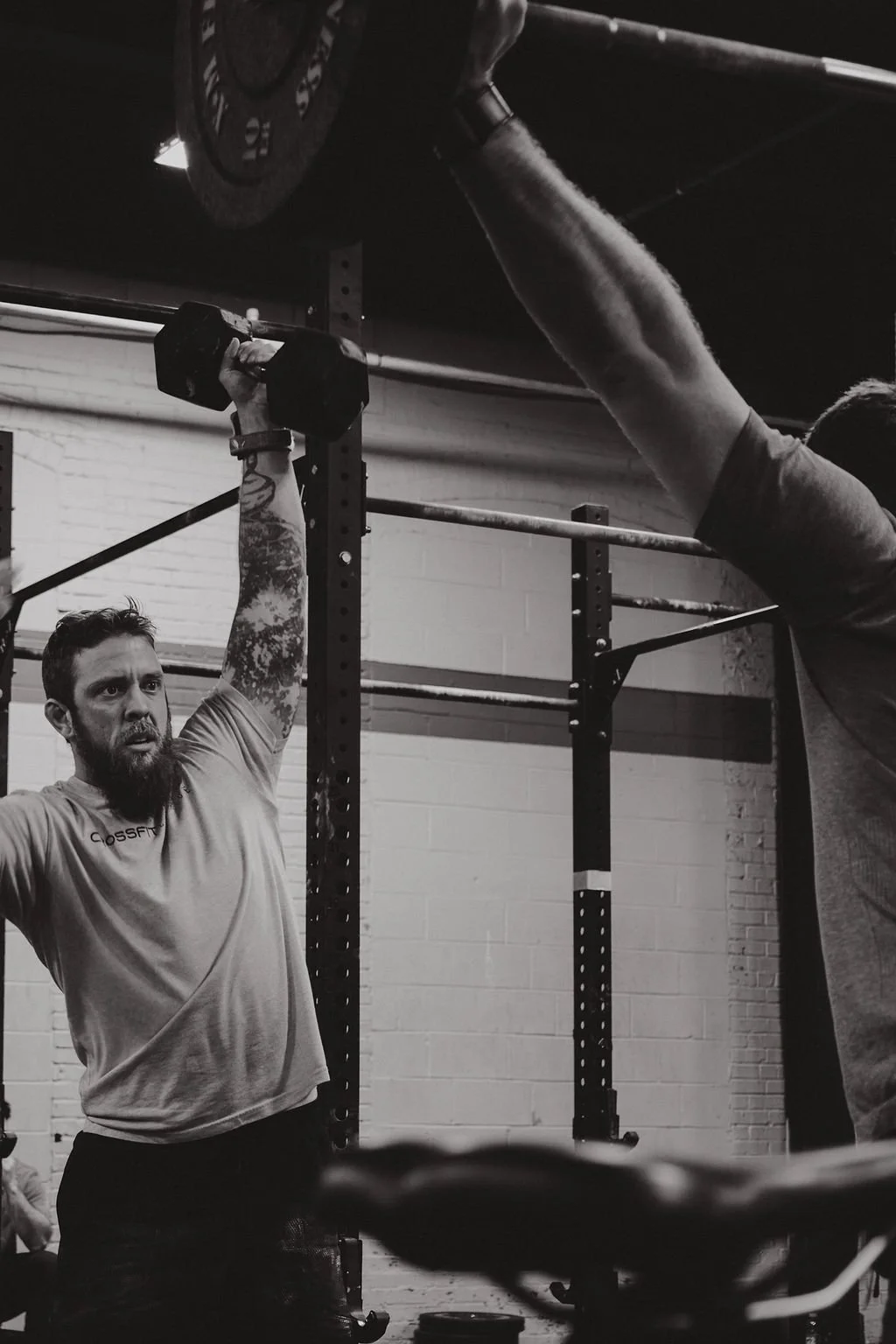 Man lifting a dumbbell overhead in a gym with a trainer nearby.
