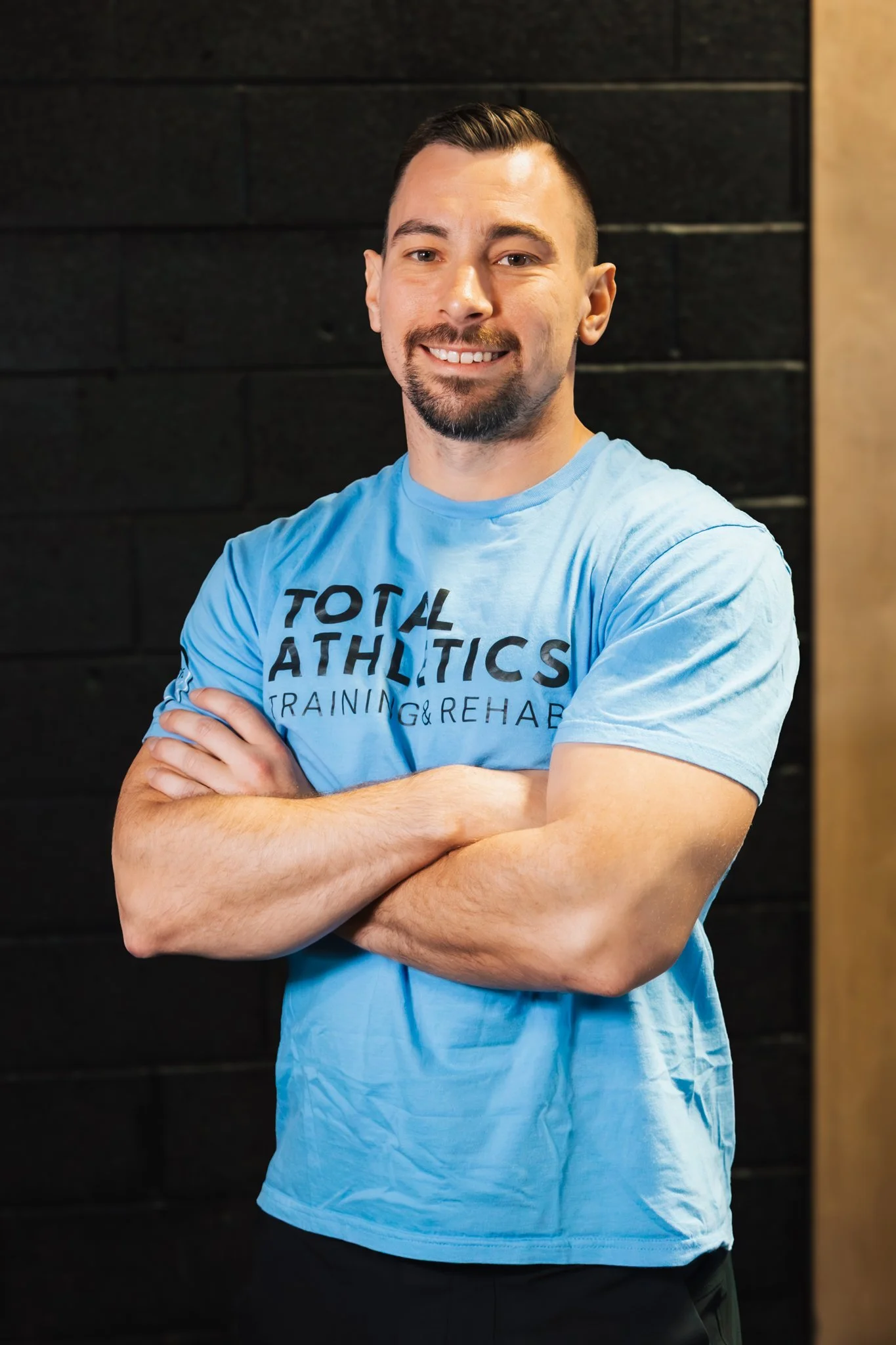 A smiling man with a goatee and short hair wearing a dark hoodie in a gym, with a folding workout bench in the background.