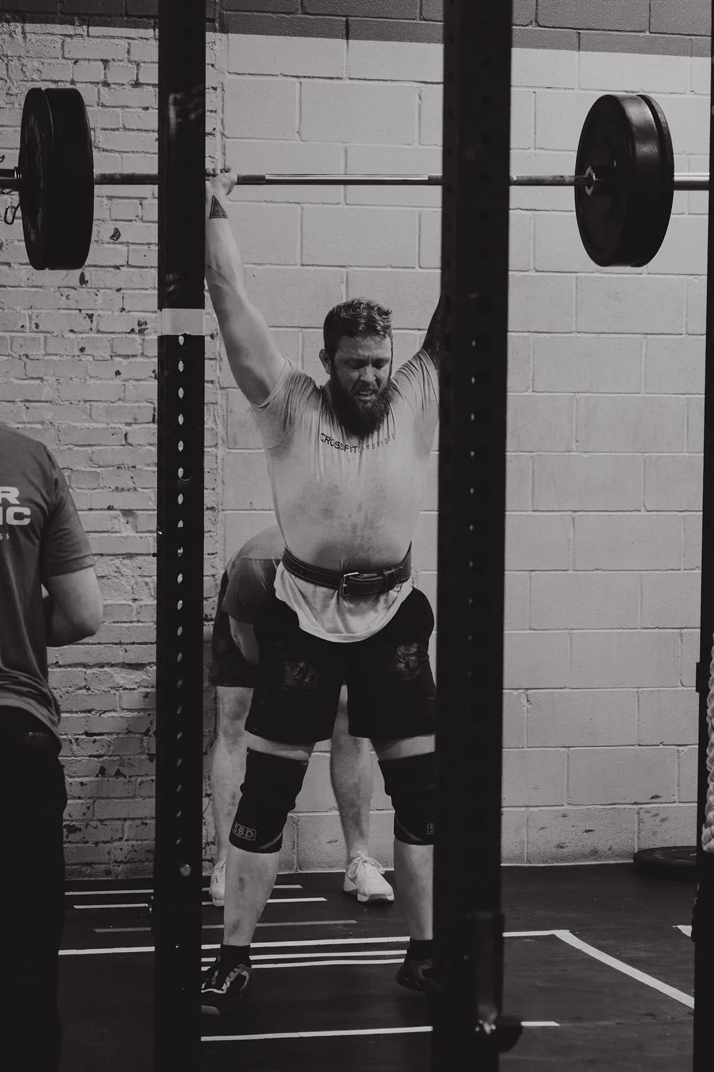 A muscular man lifting a barbell overhead in a gym, while wearing a weightlifting belt and knee sleeves, with a brick wall in the background.