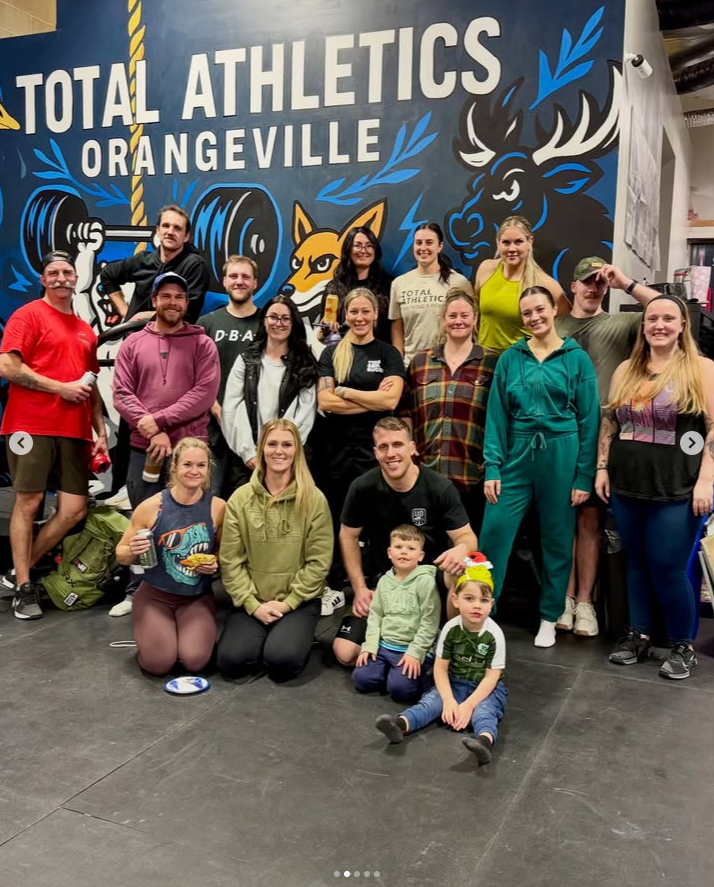 Group of people, including children, posing in front of a mural with the words 'Total Athletics Orangeville' and athletic-themed artwork in a gym or fitness center.
