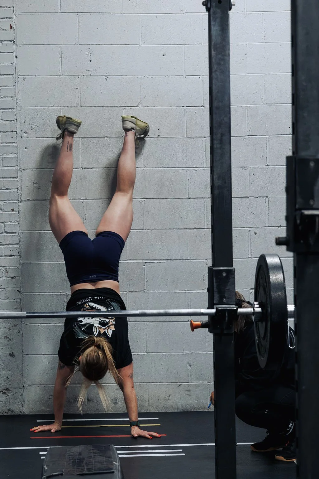 A woman doing a handstand against a gym wall with her legs extended vertically, wearing a black T-shirt with a graphic, blue shorts, and athletic shoes, in a fitness gym with weightlifting equipment.