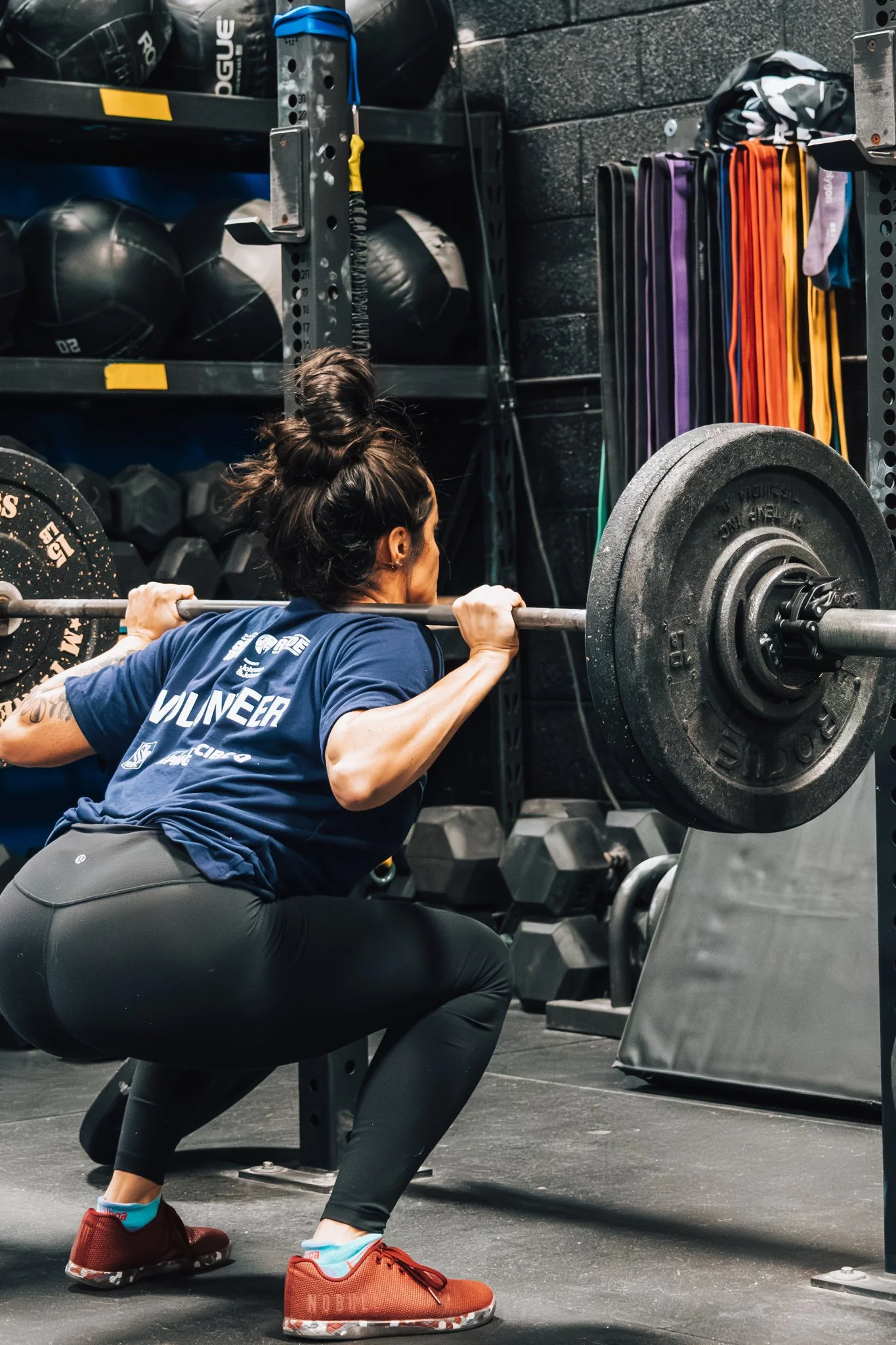 A woman with dark hair tied up, wearing a blue shirt and black leggings, is squatting while lifting a barbell with weights in a gym.