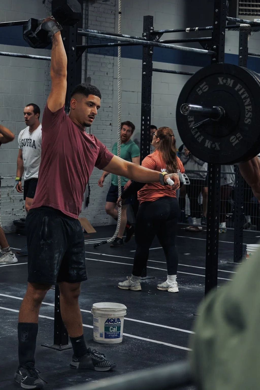 A man in a maroon shirt and black shorts is lifting a barbell overhead at a gym. Several other people are in the background, some watching, others preparing for their workout. The gym has a black floor, white brick walls, and various fitness equipmen