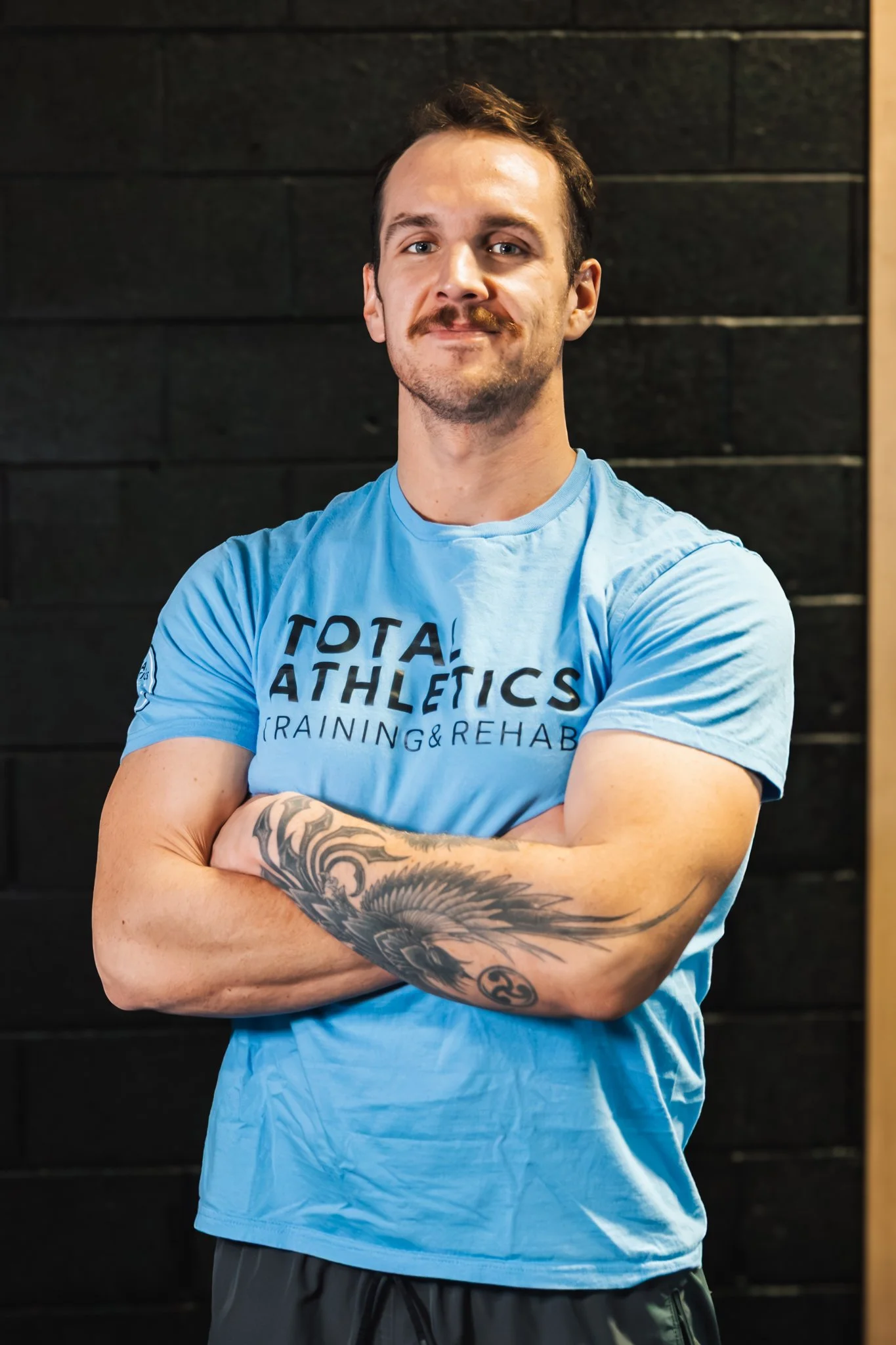 A young man in an indoor gym standing with arms crossed, smiling, wearing a beige backwards cap, black t-shirt, and green shorts, with tattoos on his right arm.