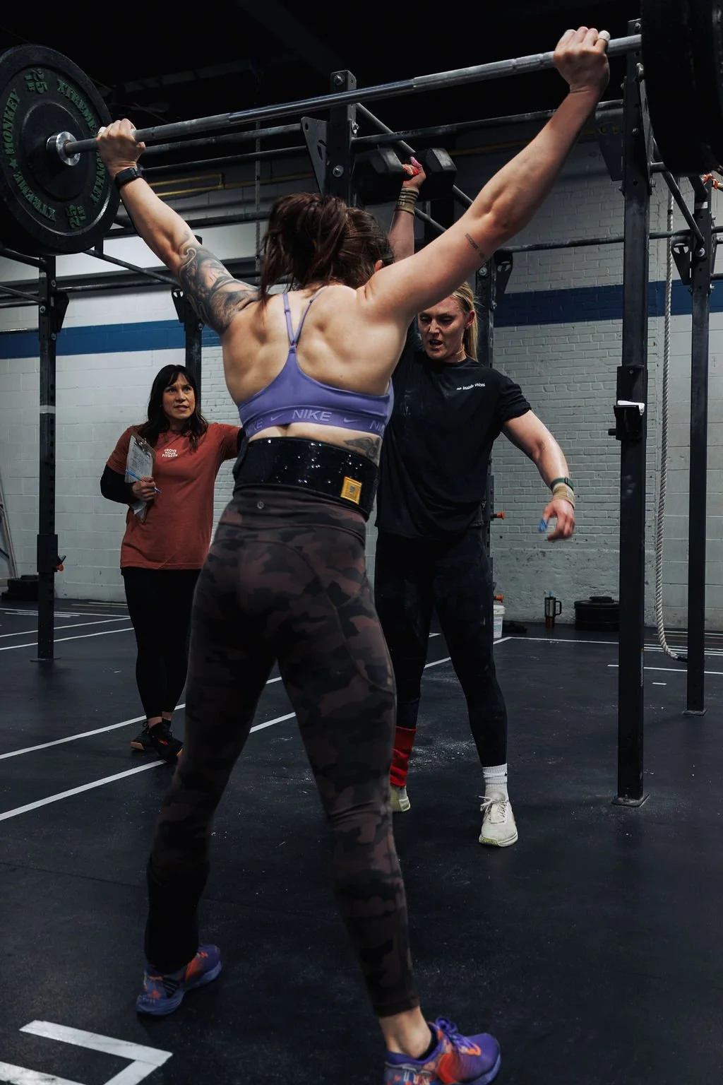 A woman with tattoos is performing an overhead barbell lift at a gym, assisted by a trainer, with another woman observing nearby.