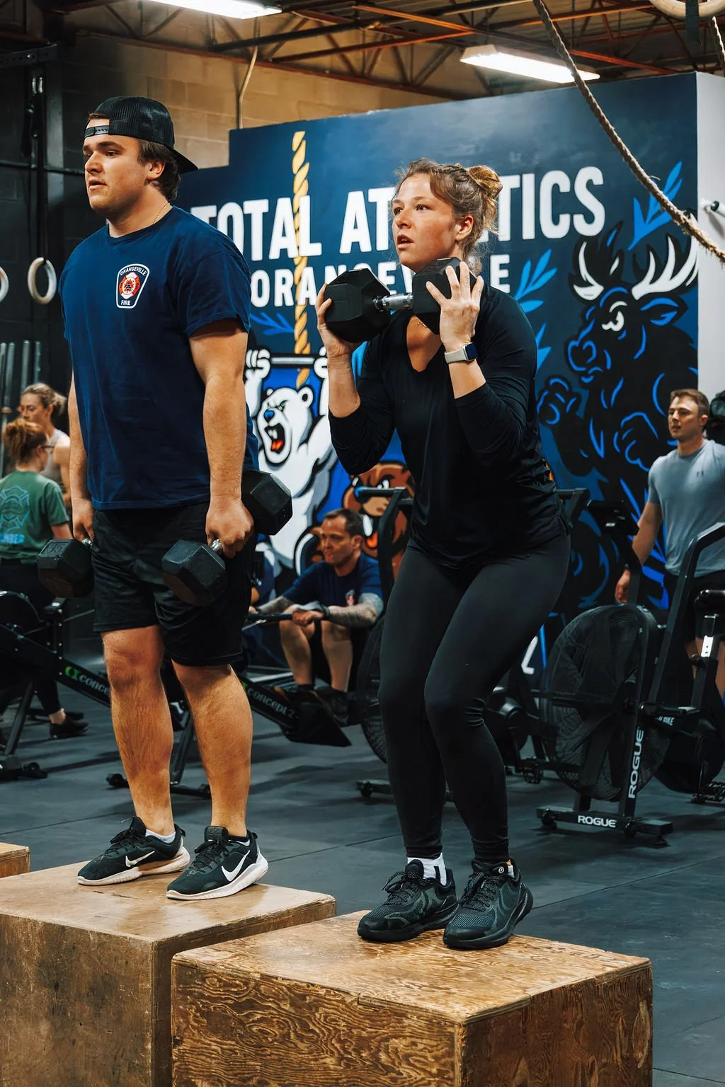 Two people lifting dumbbells on wooden platforms during a workout at an indoor gym, with a large blue banner reading 'TOTAL ATHLETICS' and people working out in the background.
