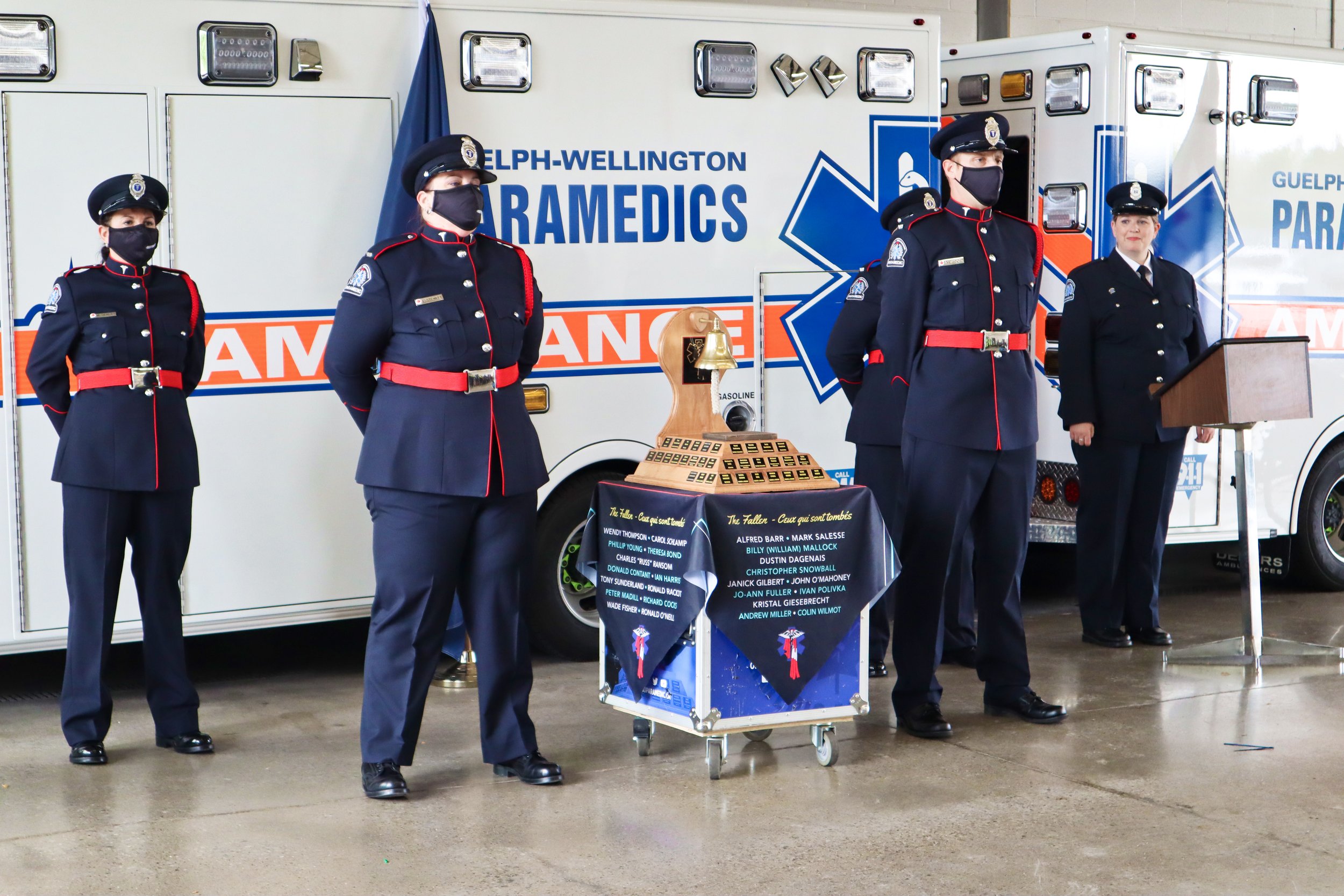 Four paramedics standing in front of an ambulance, with a memorial table and plaque in the center.