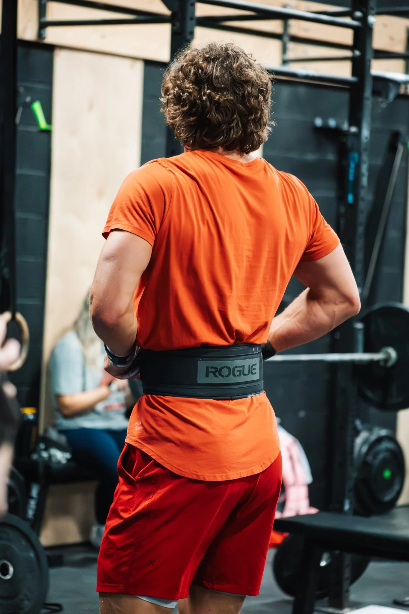 A person wearing an orange shirt and red shorts with a black Rogue weightlifting belt, standing in a gym with gym equipment and a seated woman in the background.