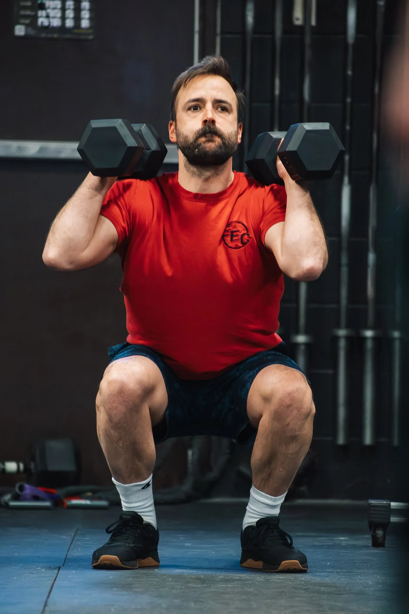 A man with a beard wearing a red shirt, black shorts, and black athletic shoes is squatting and holding dumbbells on his shoulders during a workout in a gym.
