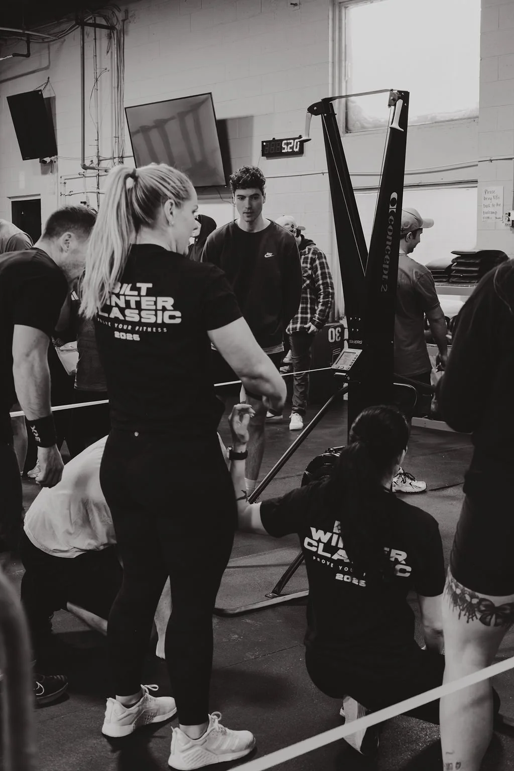 People in a gym, some preparing for a weightlifting competition, with a woman in the foreground wearing a t-shirt that says 'Built Winter Classic 2022'.