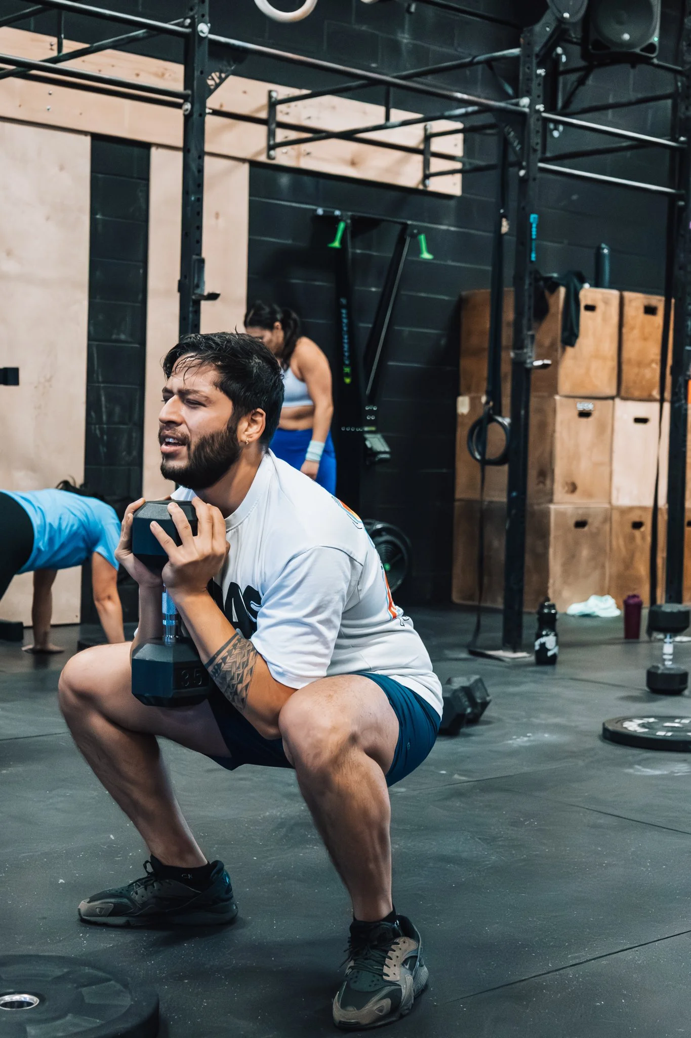 A man in a gym is holding a dumbbell close to his chest while squatting, with a focused expression on his face.