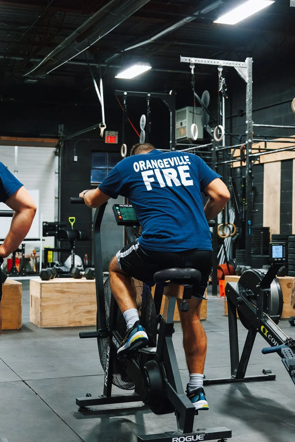 A person wearing a blue shirt with 'Orangeville Fire' on the back is exercising on a stationary bike inside a gym.