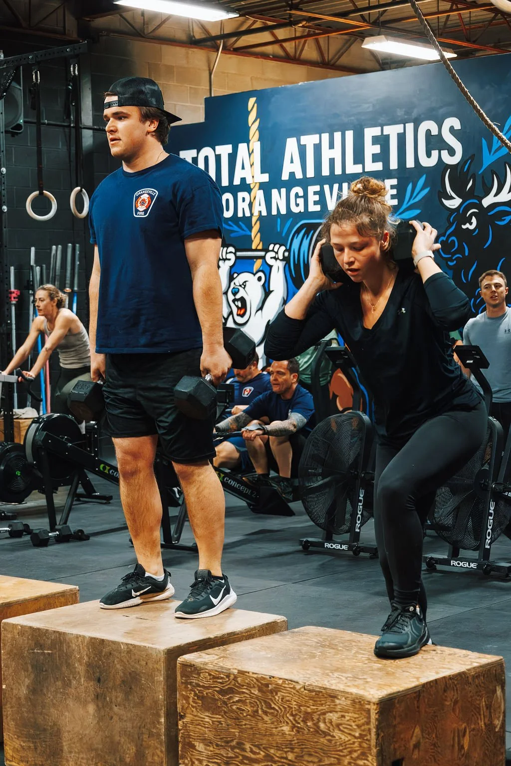 A man and a woman working out at a CrossFit gym, with the man holding dumbbells and the woman wearing a hoodie, both standing on wooden boxes. There are other people in the background, and a large sign that reads 'TOTAL ATHLETICS ORANGEVILLE' with a 
