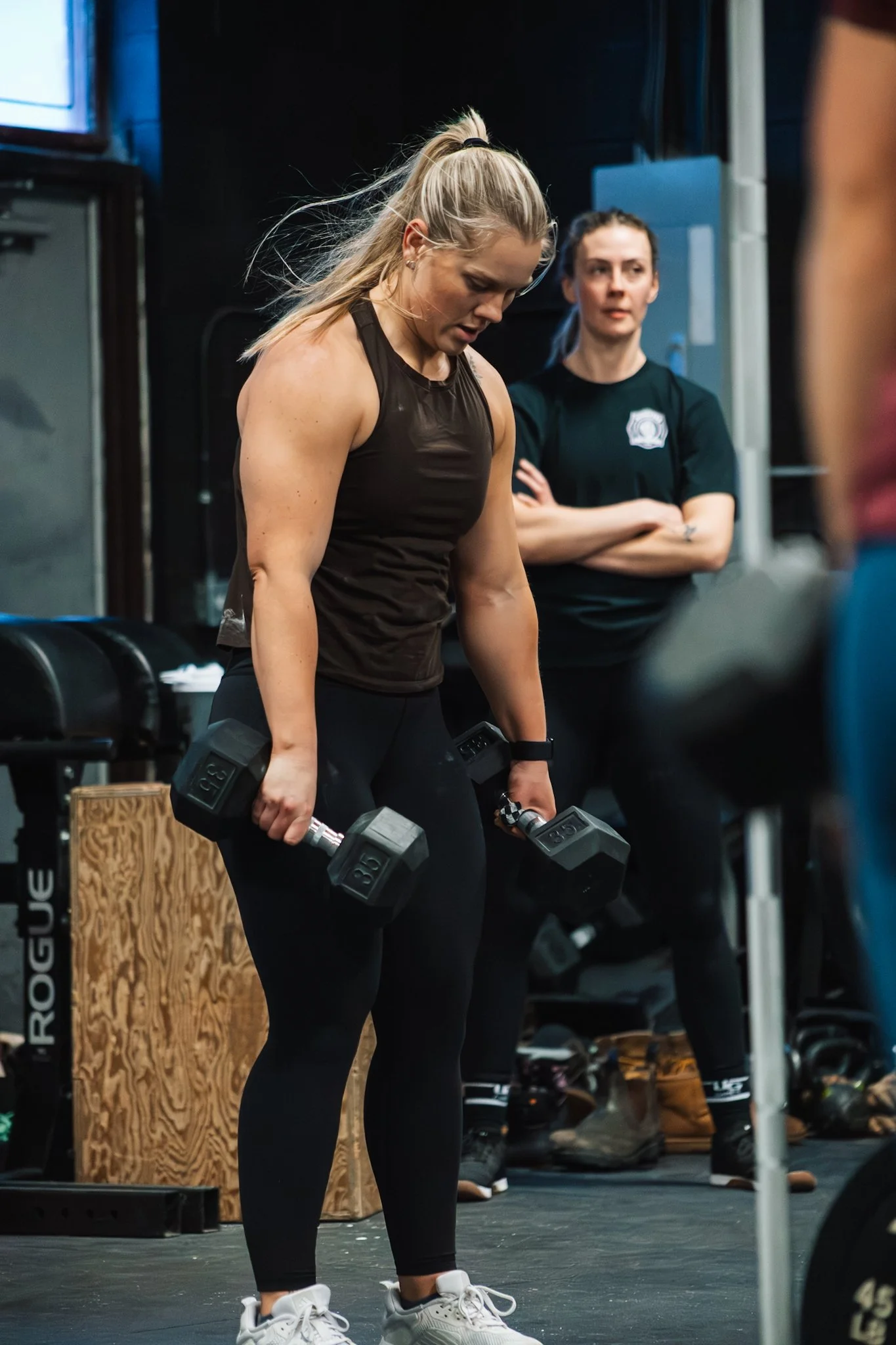 A woman in athletic clothing lifting dumbbells in a gym with an observer in the background.