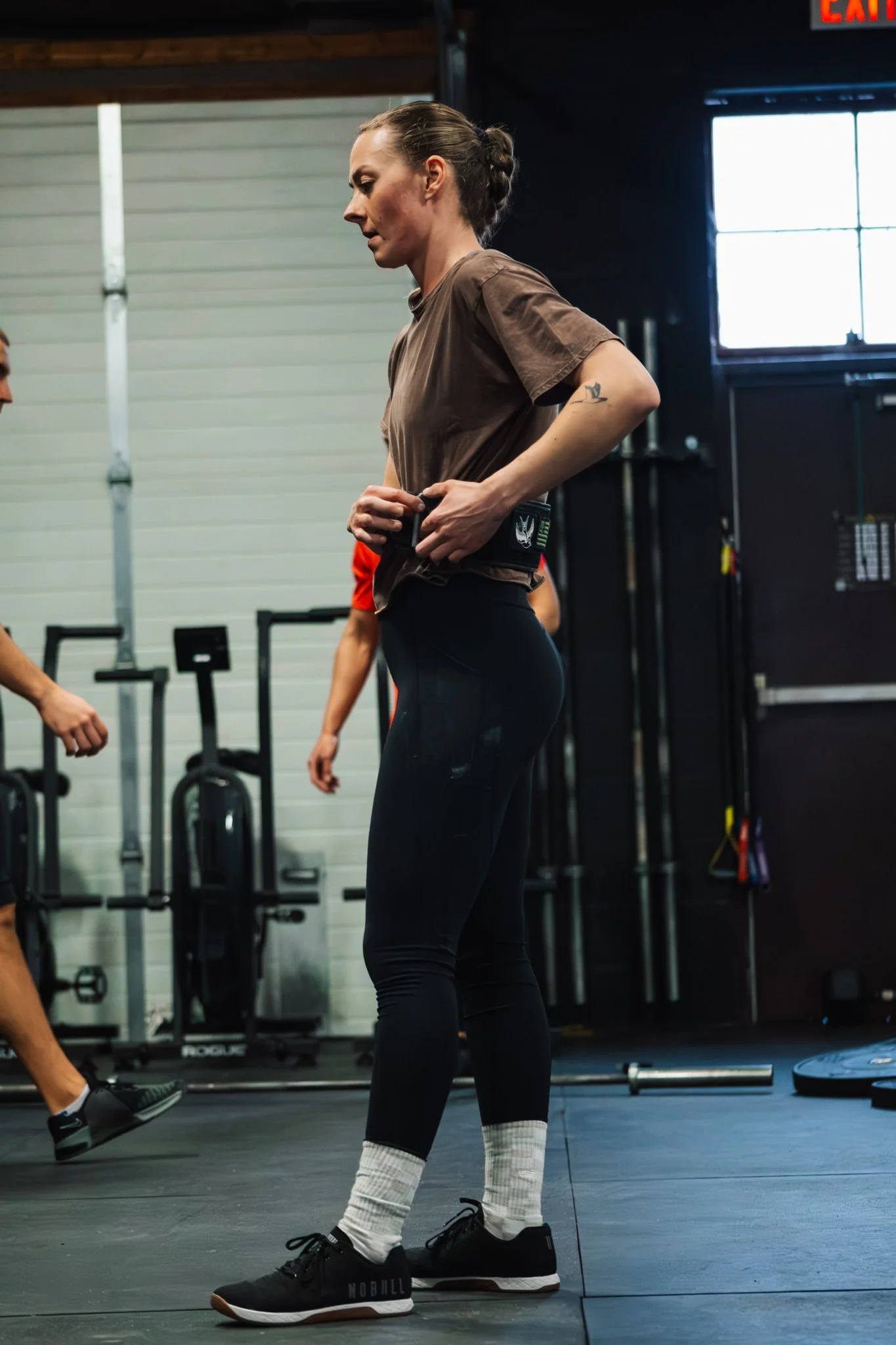 A woman standing in a gym, adjusting her weight belt. She is dressed in black leggings, a brown t-shirt, black Nike shoes, and white socks with a black stripe.