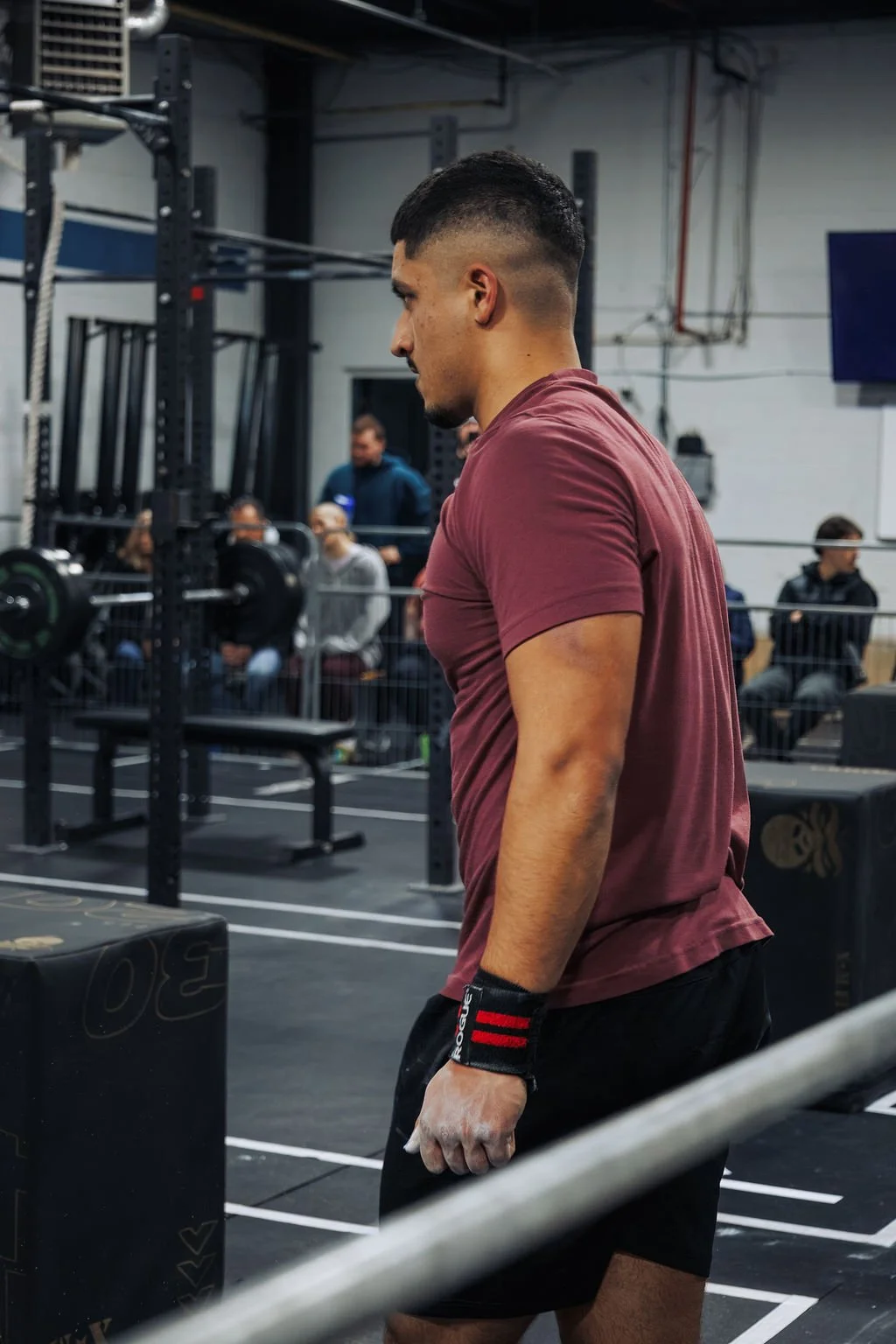 A man with a short haircut, wearing a maroon t-shirt, black shorts, and a black wristband with red stripes, stands in a gym near heavy weights and equipment, with several people seated and watching in the background.