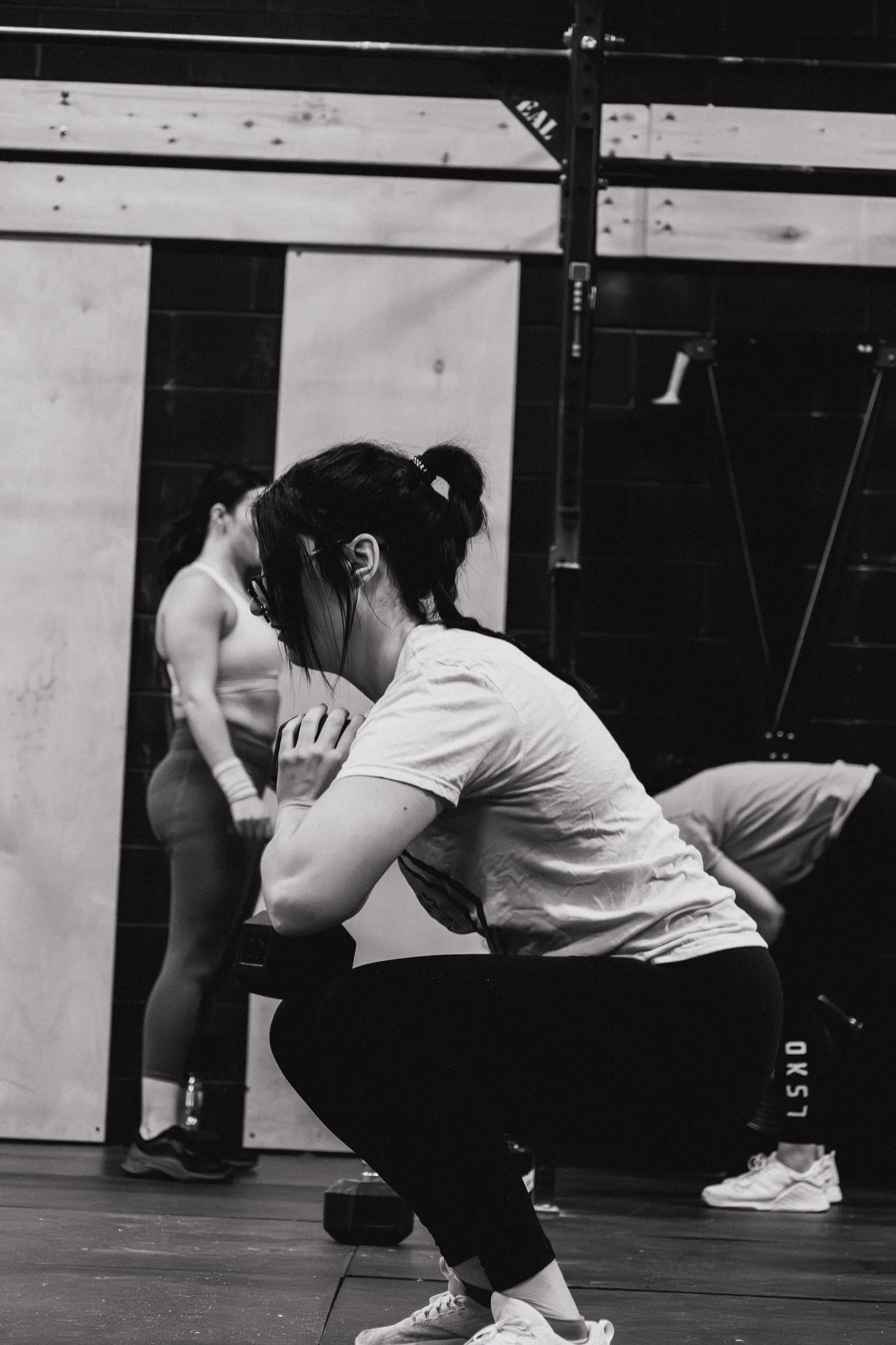A woman in workout clothing squatting in a gym with her hands clasped near her face, while another woman stands in the background watching.