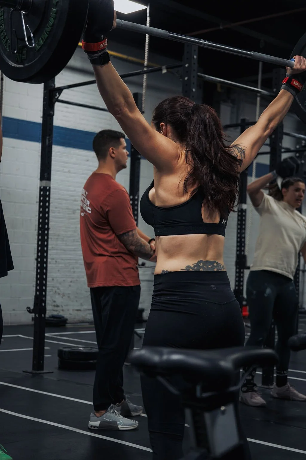 A woman performing an overhead barbell lift in a gym with other people around.