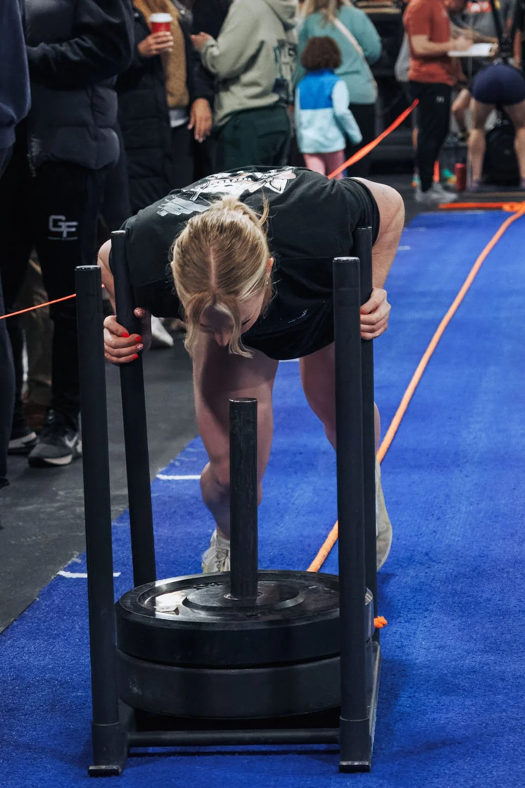 Woman pushing a heavy weight sled at a gym or fitness competition in a crowded area with onlookers and other participants in the background.