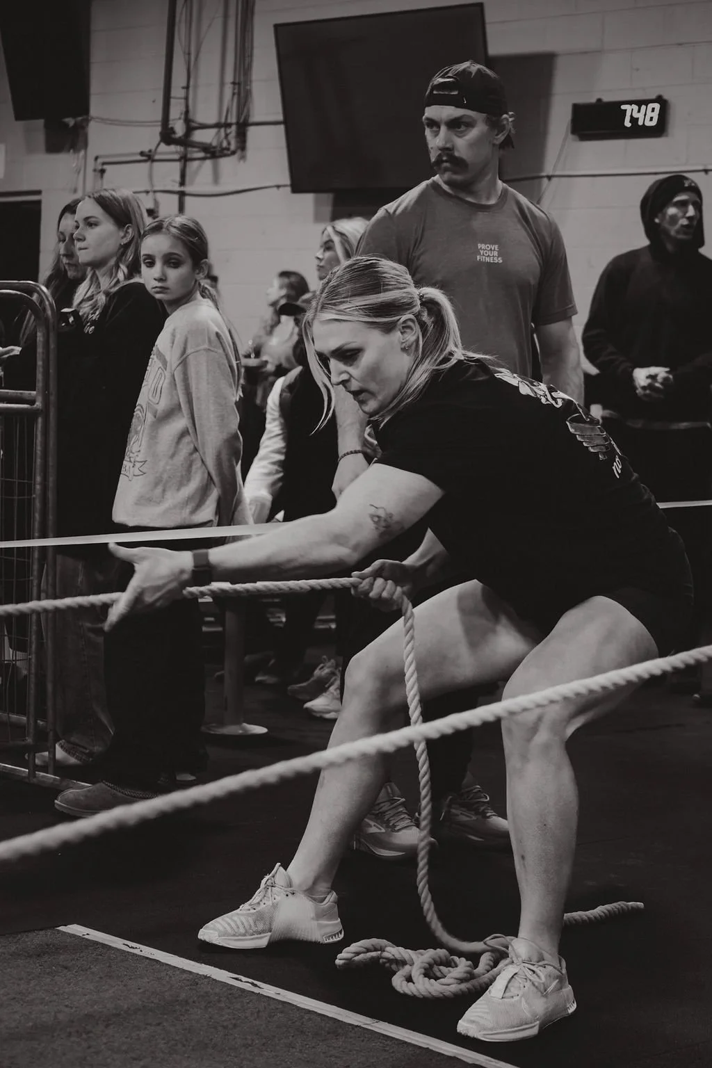 A woman participates in a tug-of-war competition inside a gym, with spectators and other participants watching. The scene is in black and white.
