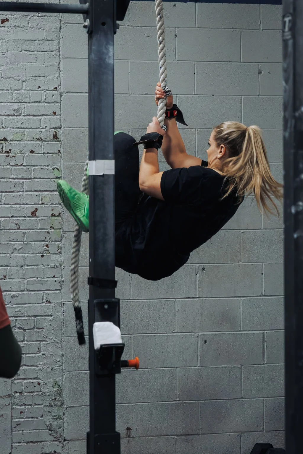 A woman with blonde hair tied in a ponytail wearing black athletic clothing and green shoes is climbing a rope inside a gym with gray cinder block walls.
