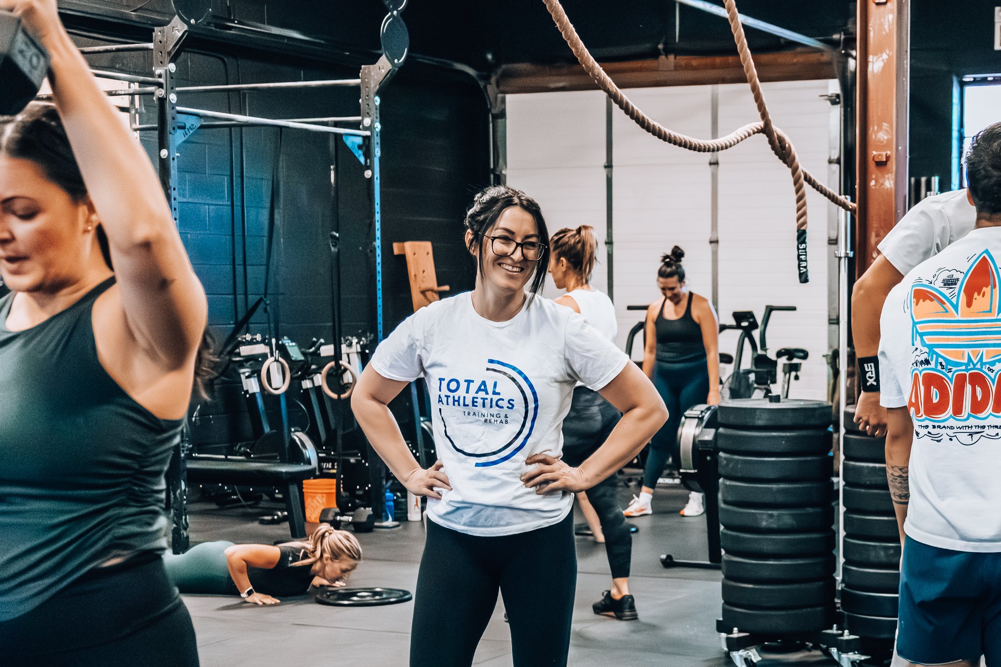 Group of women exercising in a gym, one woman in the center smiling, wearing glasses and a white 'Total Athletics' T-shirt, surrounded by workout equipment and other women exercising.