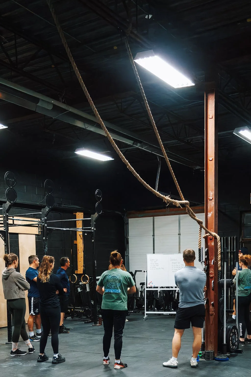 Group fitness class in a gym with a whiteboard, fitness equipment, and a climbing rope hanging from the ceiling.