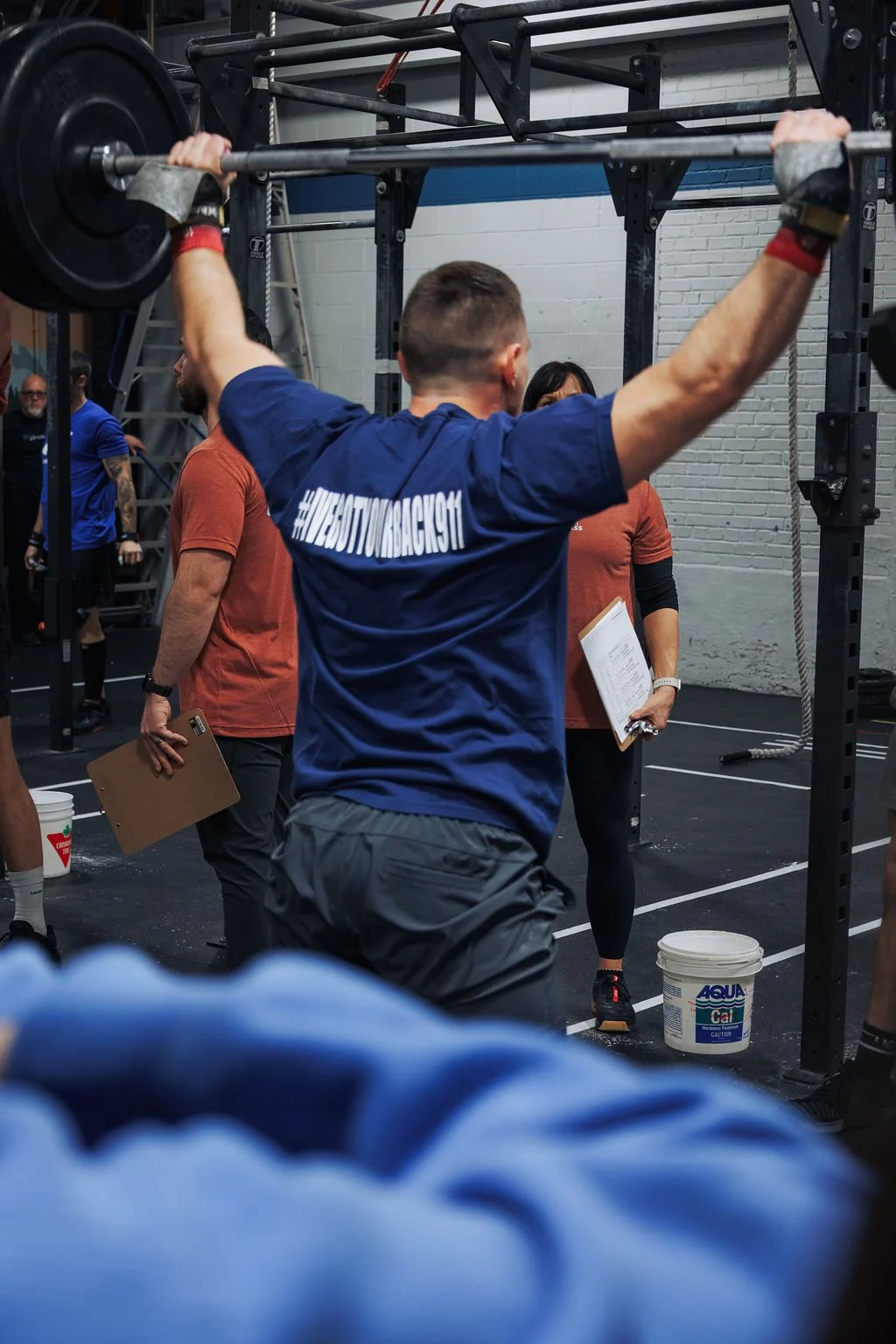 Man lifting a barbell at a CrossFit gym with other people in the background, holding clipboards and wearing workout gear.