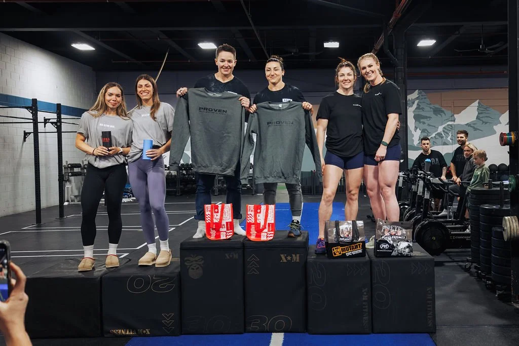 Group of women standing on a podium at a gym, holding prizes and shirts after a competition or event.