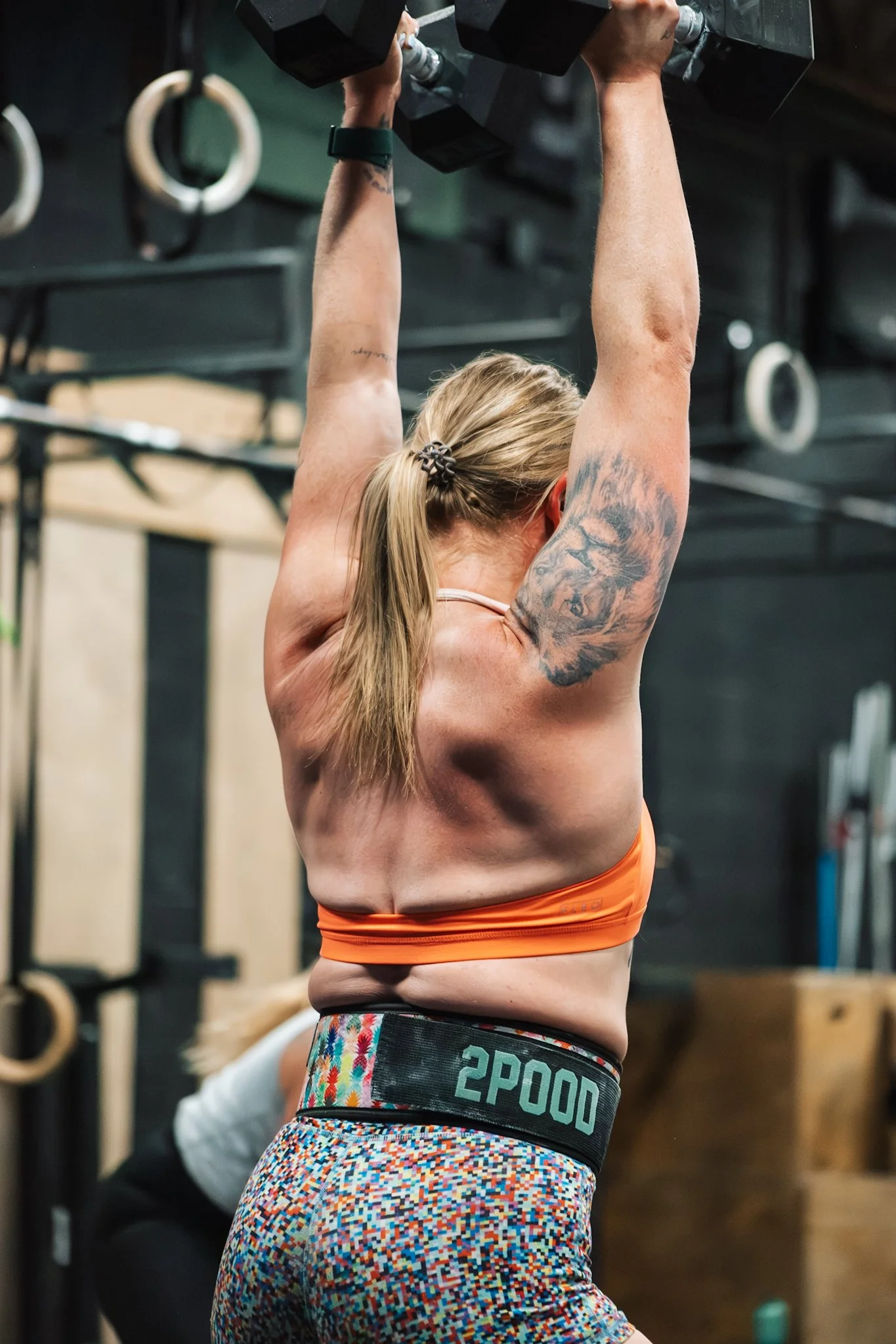 A woman lifting dumbbells overhead in a gym, wearing a sports bra and colorful leggings.