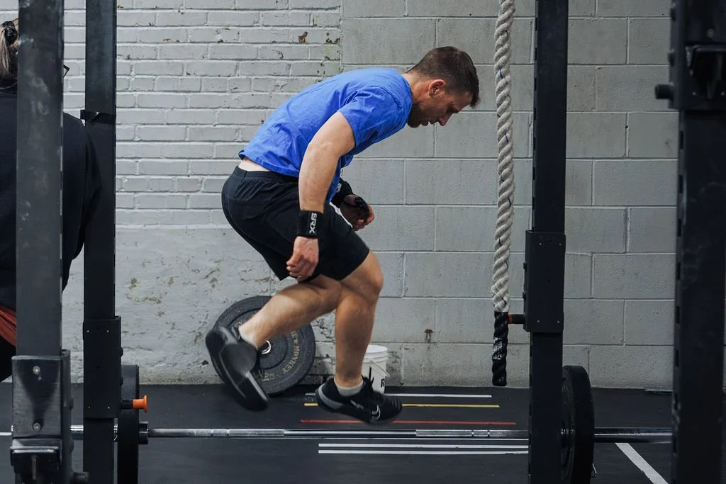 Man is mid-air, jumping inside a gym near a barbell on the floor.