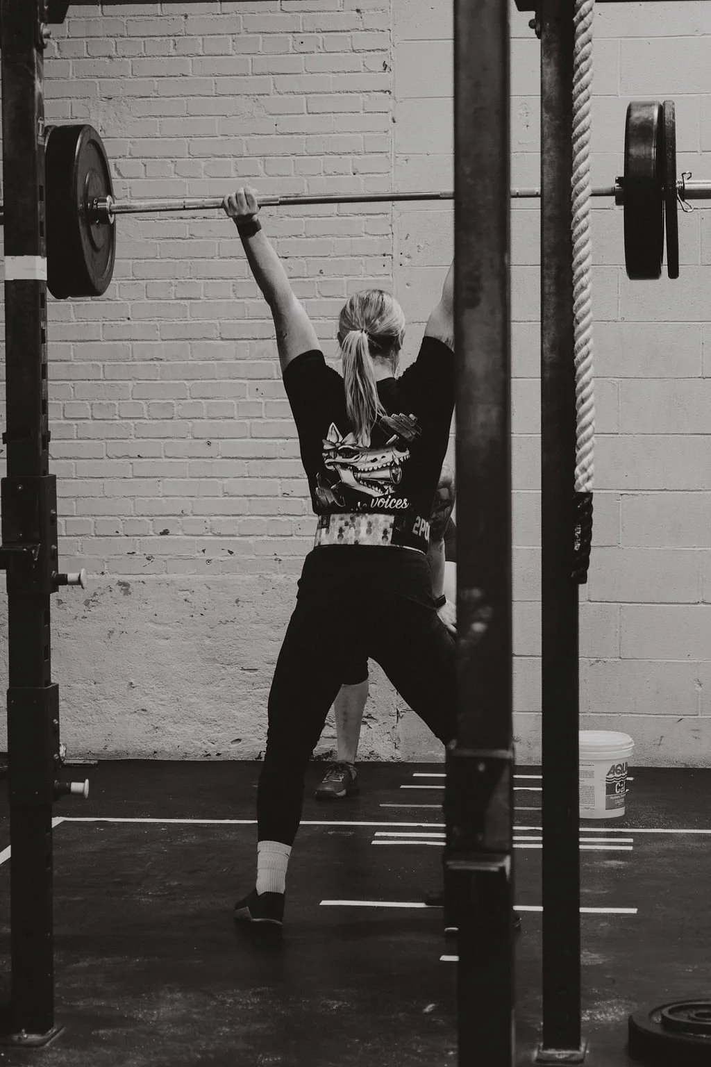 A woman lifting a barbell during a workout in a gym with a brick wall background, black and white photo.
