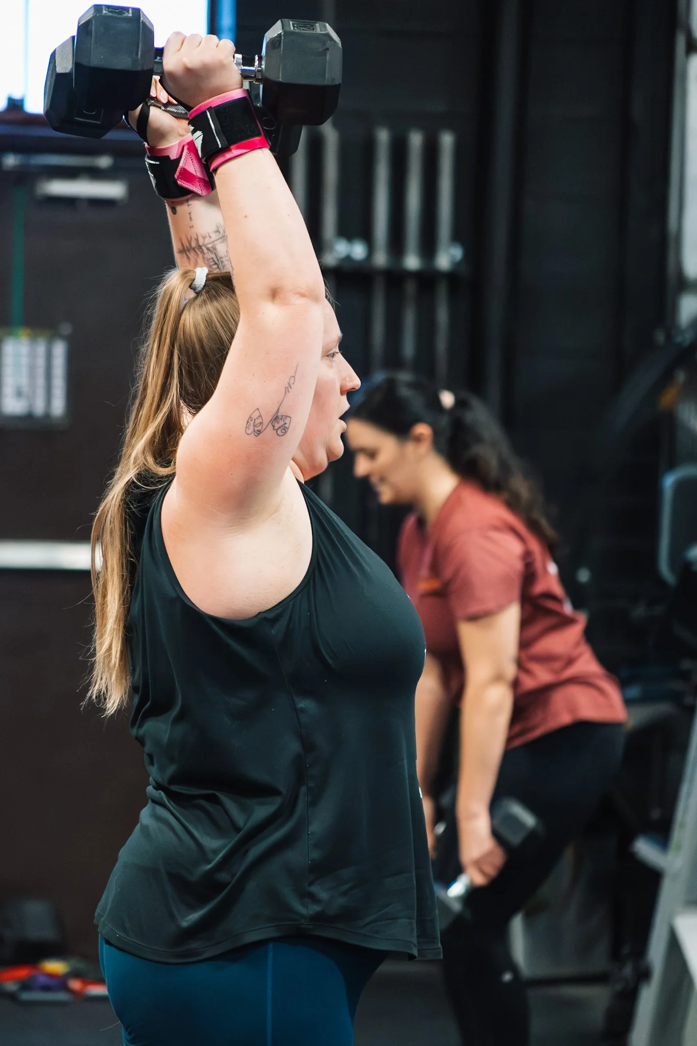 A woman lifting a dumbbell overhead during a workout at the gym, with another woman in the background also exercising.