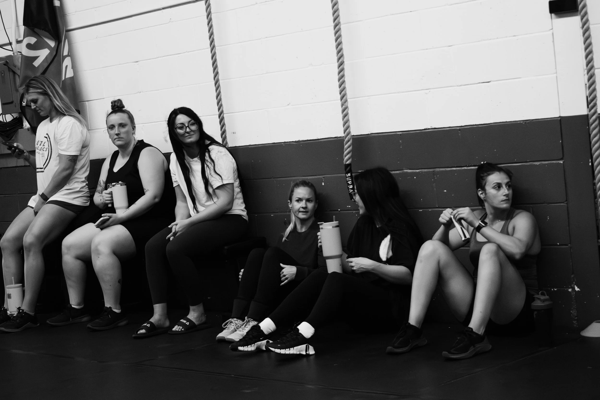 Six women sitting on the floor against a wall, some holding water bottles, in a gym or sports facility.
