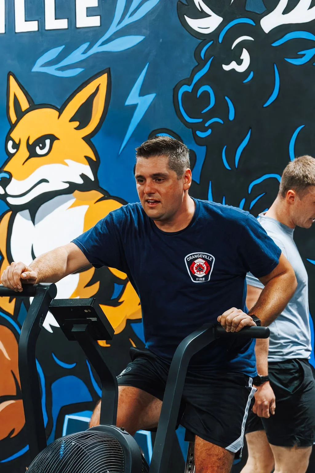 Man working out on a stationary bike in a gym with a colorful mural of animals, including a fox and a deer, in the background.