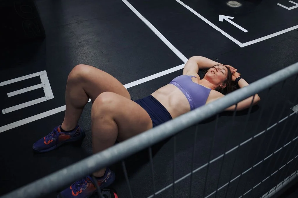 A woman lying on the ground in a parking lot after exercise, wearing a purple sports bra and shorts, with her eyes closed and arms resting behind her head.