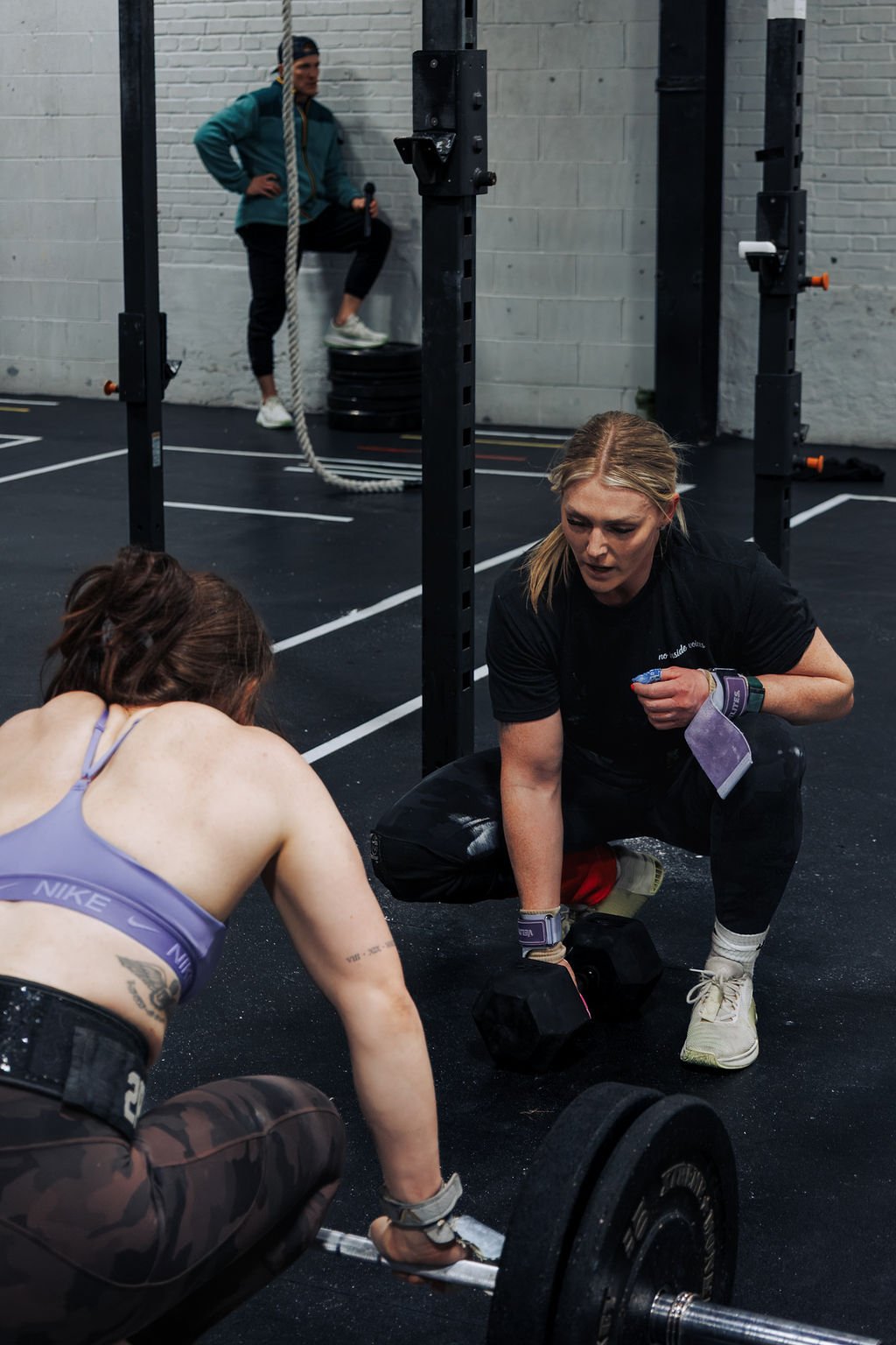 Two women are doing a workout on a gym floor with weights, while a person in the background is lifting a heavy bag and holding a microphone.